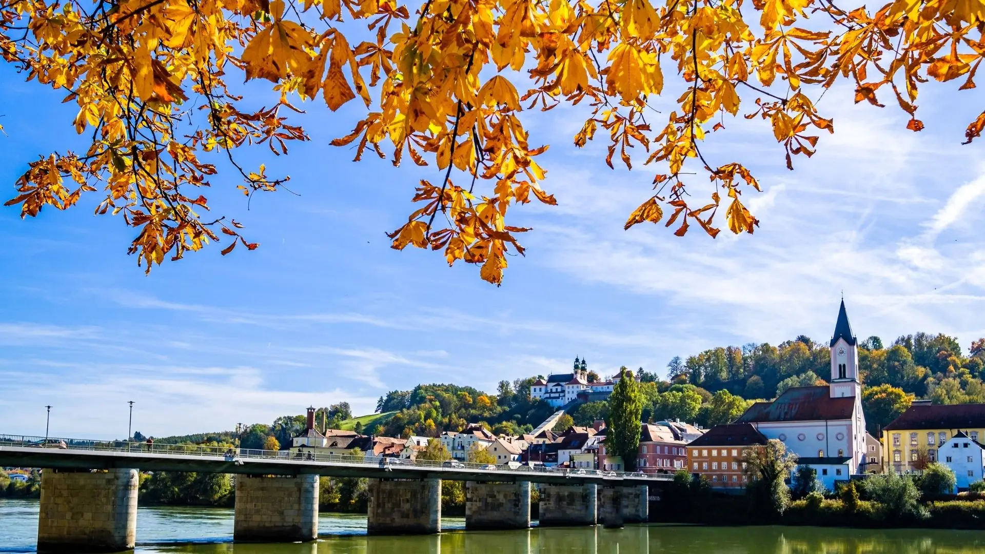 Brücke über den Fluss mit mehreren Bögen, umgeben von herbstlichen Bäumen mit gelben Blättern und einer Stadtansicht im Hintergrund.