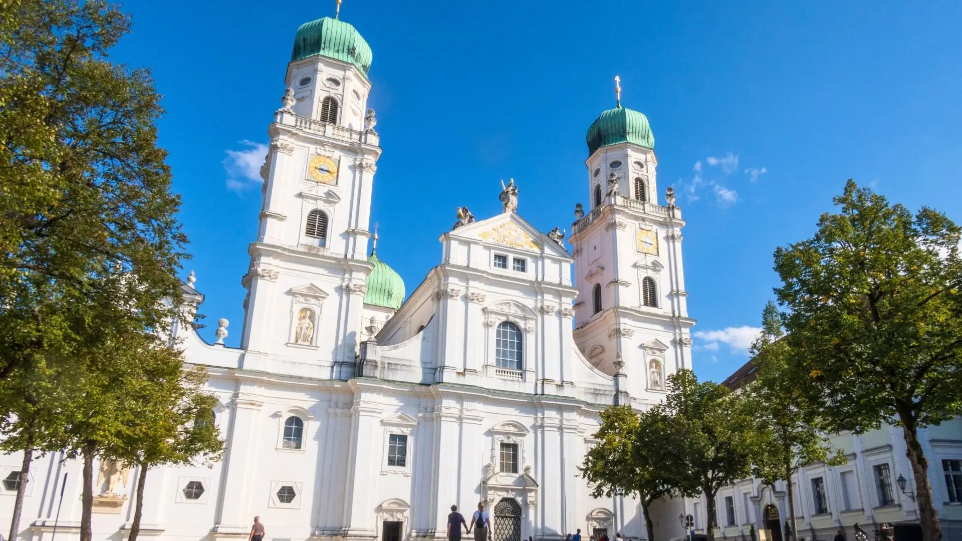 Die Kathedrale St. Stephan in Passau mit zwei hohen Türmen und grünen Kuppeln, umgeben von Bäumen und einem blauen Himmel.