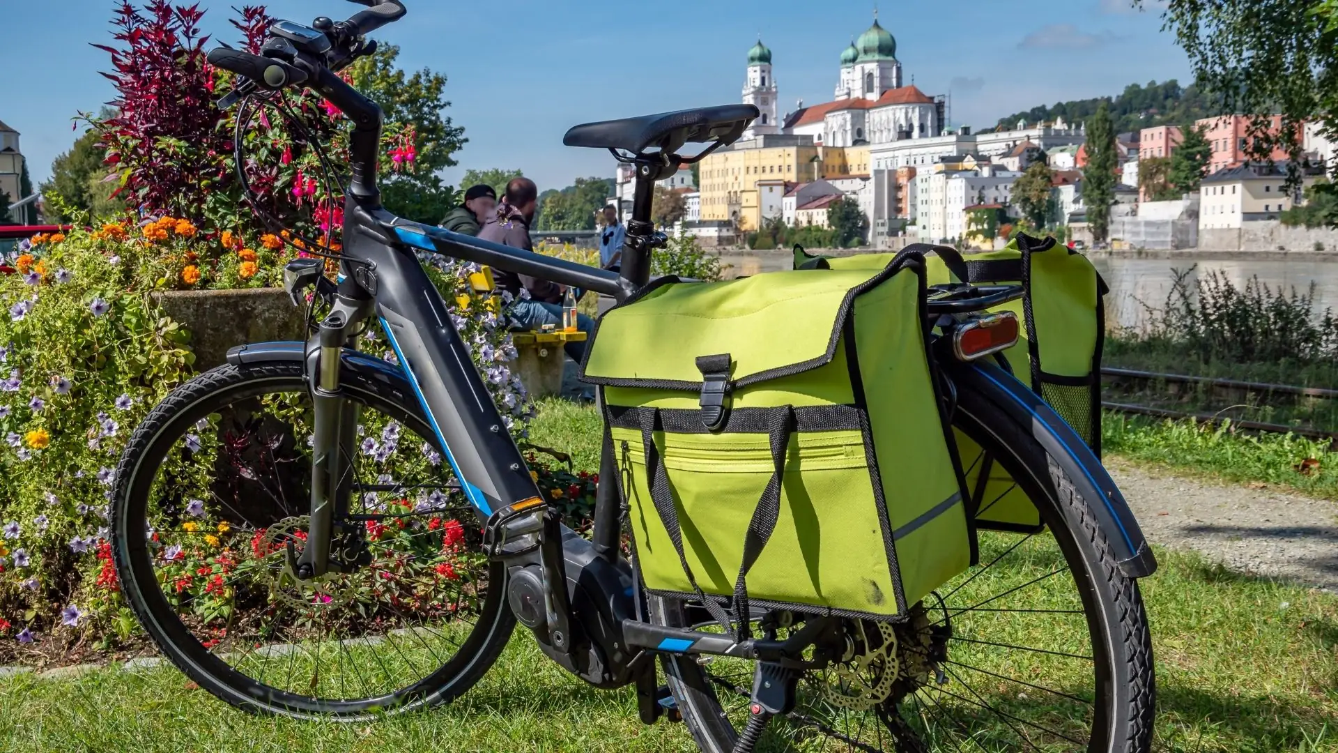 Schwarzes E-Bike mit zwei grünen Gepäcktaschen, im Vordergrund einer blühenden Blumenlandschaft, im Hintergrund die Stadt Passau mit historischen Gebäuden.