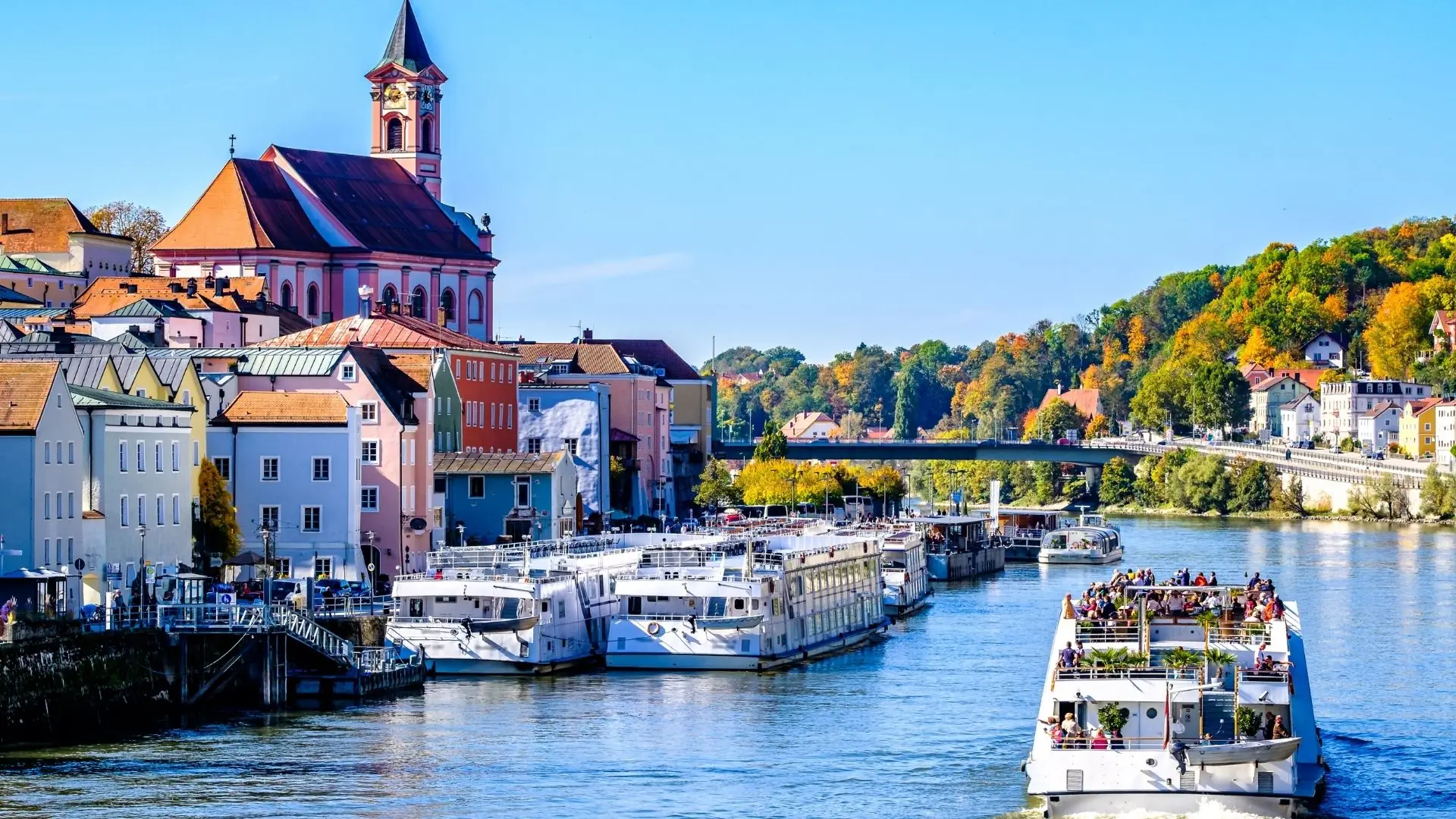 Fluss mit mehreren Booten im Vordergrund, im Hintergrund farbenfrohe Gebäude und eine Kirche mit einem hohen Turm, Bäume mit herbstlichen Farben.