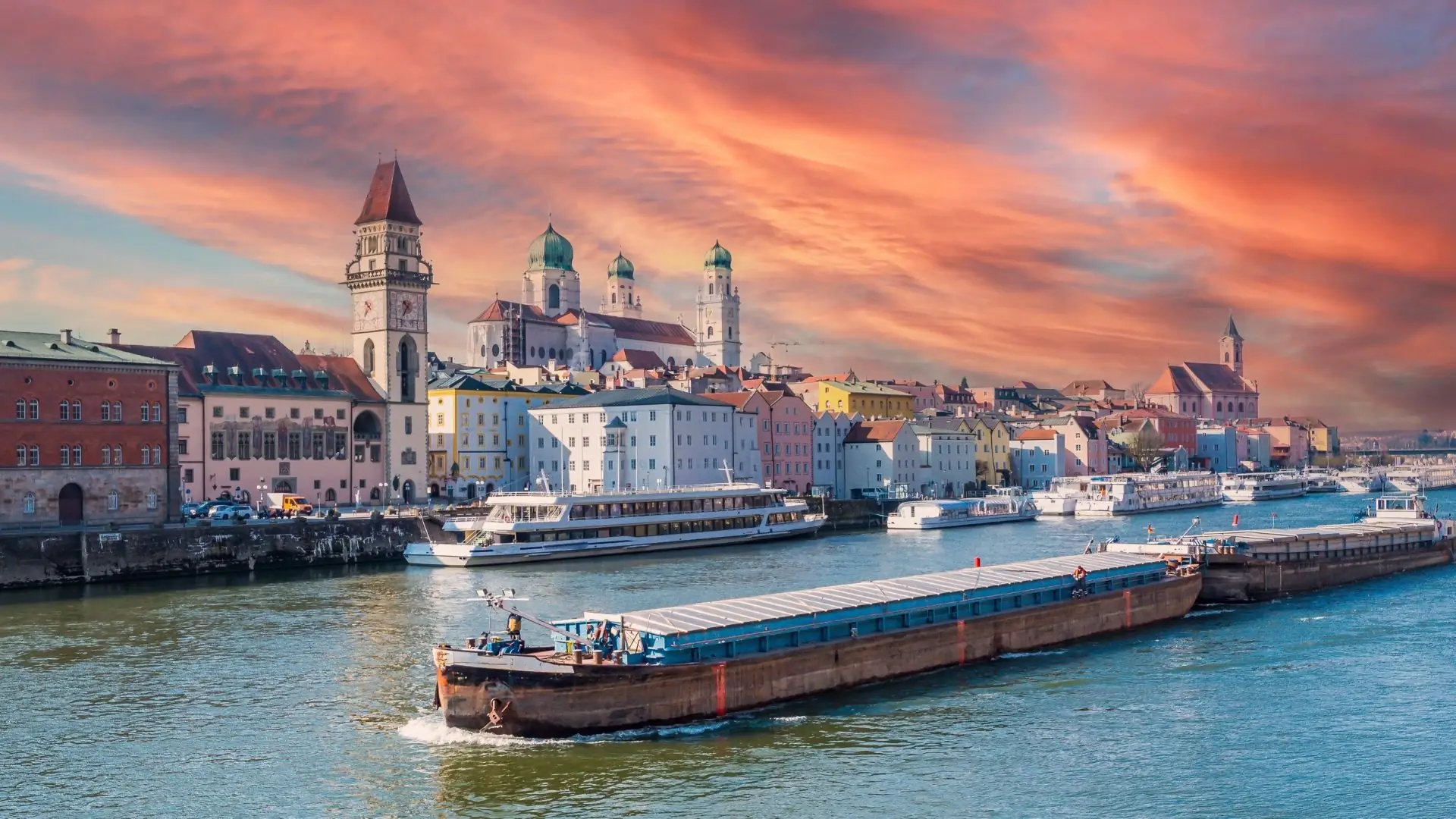 Blick auf die Stadt Passau mit bunten Gebäuden am Ufer, einem Frachtschiff auf dem Wasser und einer dramatischen Wolkenlandschaft im Hintergrund.