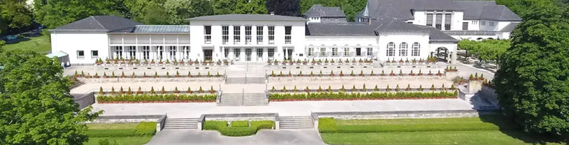 Luftaufnahme des Dolce Bad Nauheim mit einer begrünten Terrasse, umgeben von üppigem Grün und bunten Blumenbeeten.