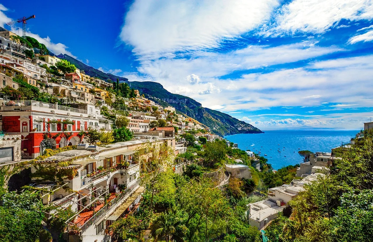Positano, mit bunten Häusern, die sich aneinanderreihen und harmonisch in die Berglandschaft eingefügt sind. Die Aussicht auf das blaue, glitzernde Meer verleiht der Szenerie eine malerische und einladende Atmosphäre, während die lebendigen Farben der Architektur im Sonnenlicht erstrahlen.