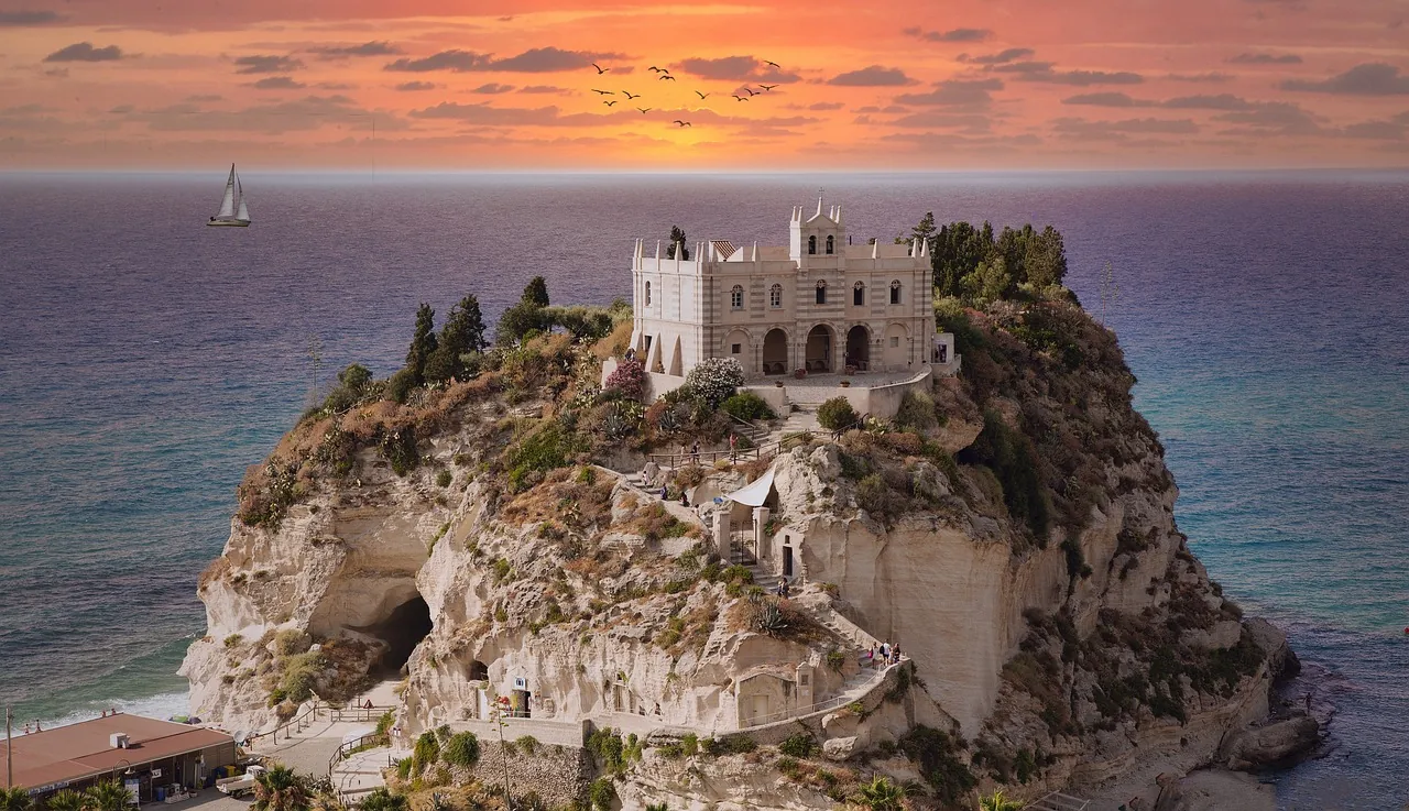 Aussicht auf die Kirche von Tropea, die malerisch auf einem hohen Hügel mit Bäumen und Pflanzen thront, während im Hintergrund ein Segelschiff auf dem glitzernden Meer fährt. Der leicht rötliche Himmel beim Sonnenuntergang wird von einer Gruppe Vögel, die am Himmel fliegen, geschmückt und schafft eine romantische und idyllische Atmosphäre.