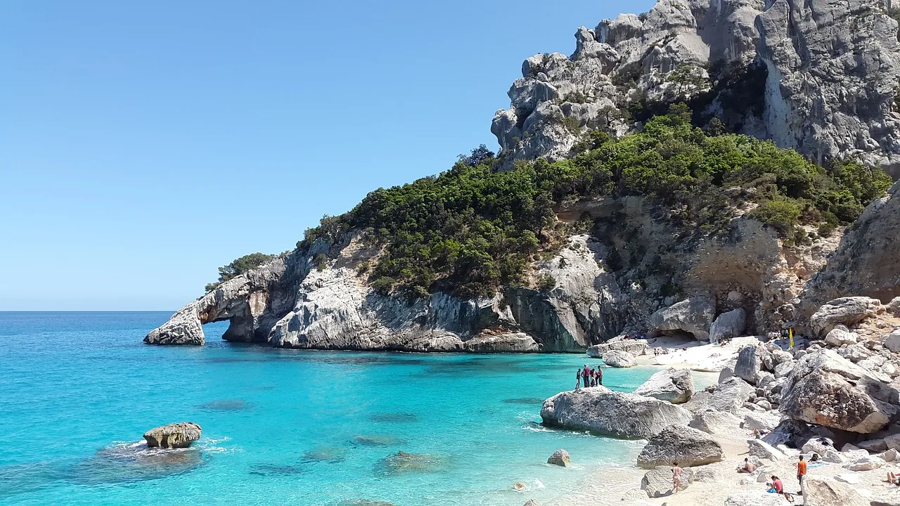 Sardinien, ein weißer Sandstrand mit hellblauem Wasser an der Küste. Einige Menschen entspannen sich in der Sonne und genießen den Blick auf die malerische Umgebung, während im Hintergrund beeindruckende Berge die Landschaft umrahmen und eine idyllische Atmosphäre schaffen.