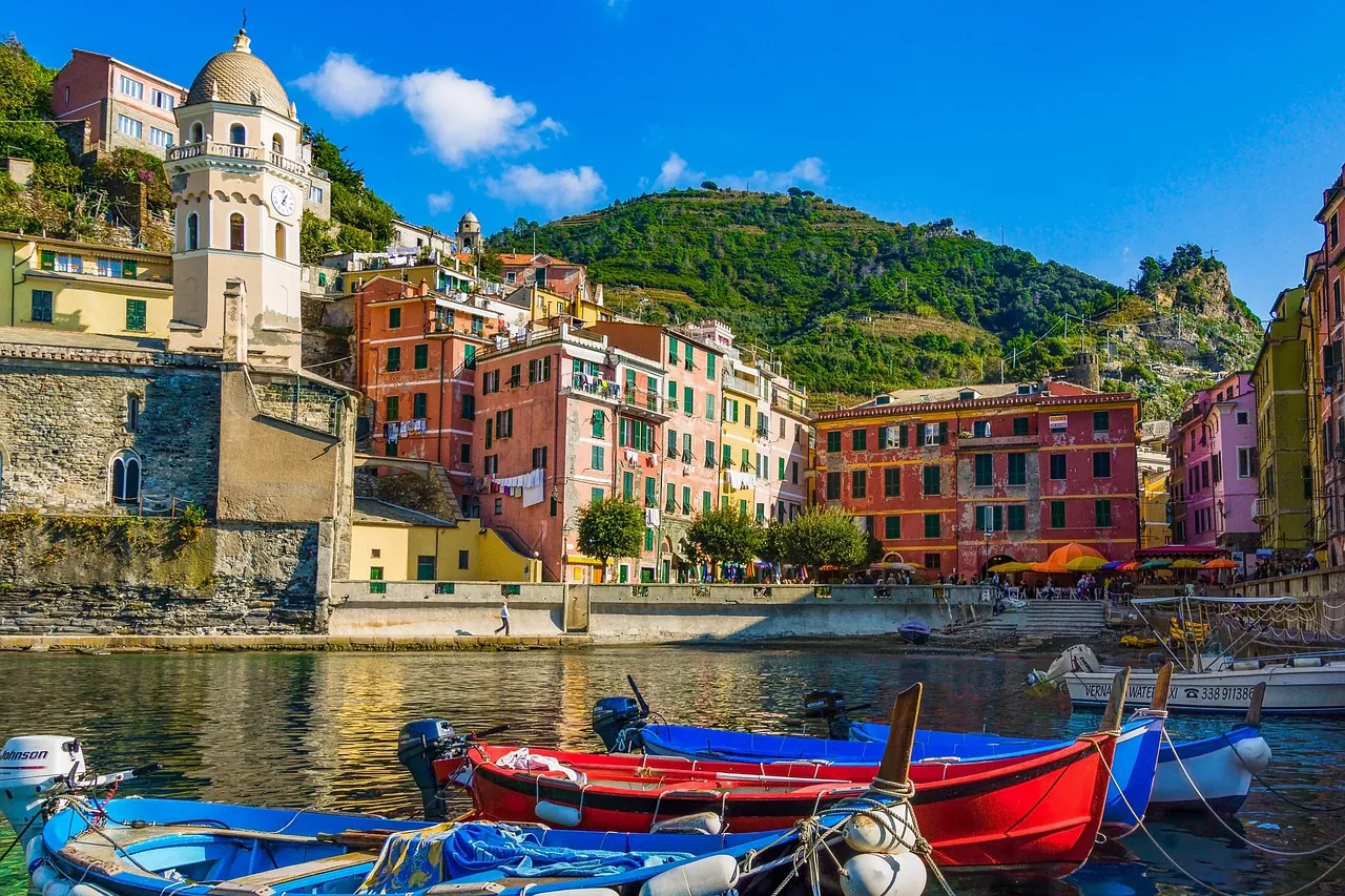 Aussicht auf den Hafen von Cinque Terre, mit farbenfrohen Häusern, die sich malerisch an die grünen Hügel schmiegen. Das Meer, gesäumt von angelegten Booten, ergänzt die lebendige Kulisse und schafft eine einladende maritime Atmosphäre.