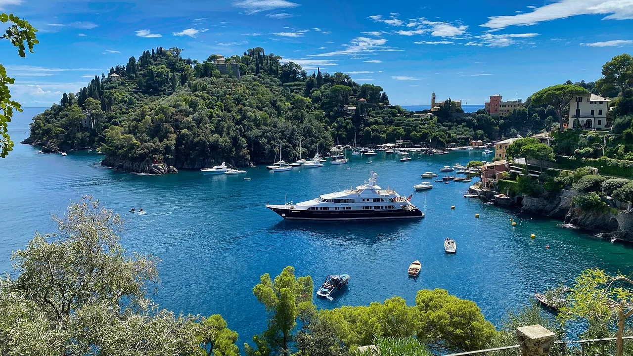 Aussicht auf den Hafen von Portofino, gesäumt von Yachten und Schiffen, eingebettet in eine atemberaubende Berglandschaft mit üppiger Vegetation. Das glitzernde Meer spiegelt das Sonnenlicht wider und verleiht der Szene eine einladende und malerische Atmosphäre.