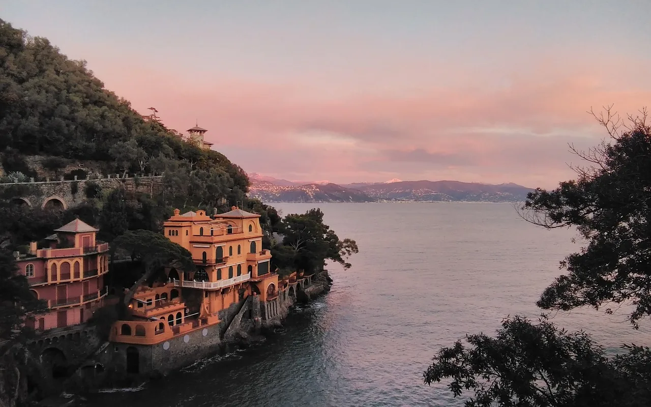 Portofino, mit bunten Häusern, die direkt über dem Meer thronen und förmlich am Berg kleben. Bei Sonnenuntergang erstrahlen die Fassaden in warmen Farben, während das Licht des Sonnenuntergangs das glitzernde Wasser und die malerische Umgebung in eine romantische Atmosphäre taucht.