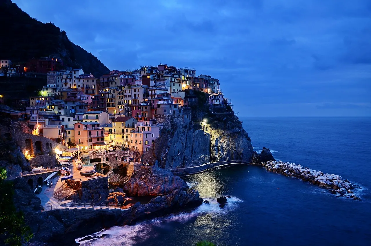 Aussicht auf Cinque Terre bei Nacht, wo die leuchtenden bunten Häuser an den Klippen eine zauberhafte Atmosphäre schaffen. Die stimmungsvolle Beleuchtung reflektiert sich im schönen ozeanblauen Meer und betont den einzigartigen Charme dieser malerischen Küstenlandschaft.