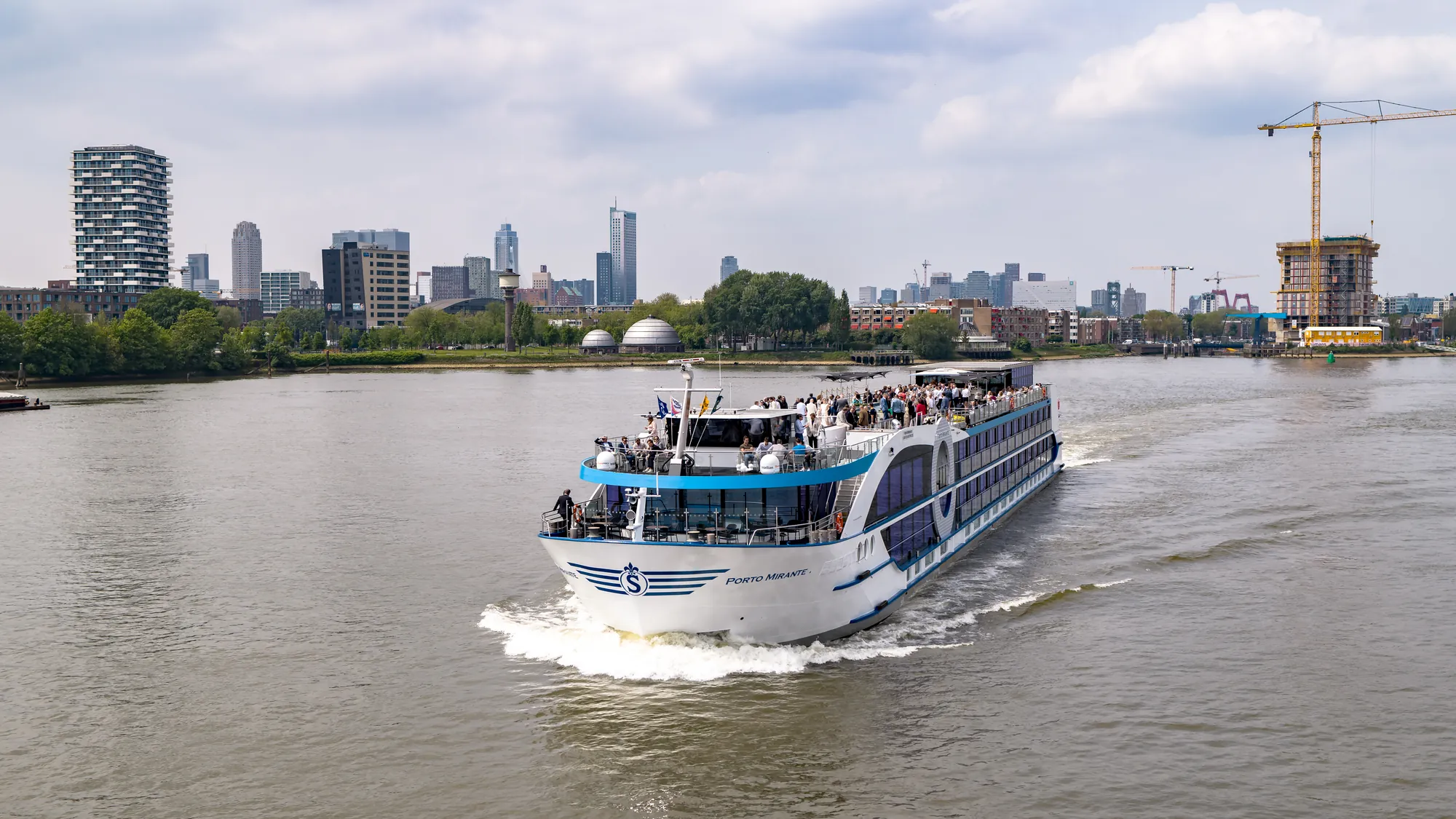 Ein Kreuzfahrtschiff fährt auf einem ruhigen Fluss mit einer Stadtsilhouette im Hintergrund.