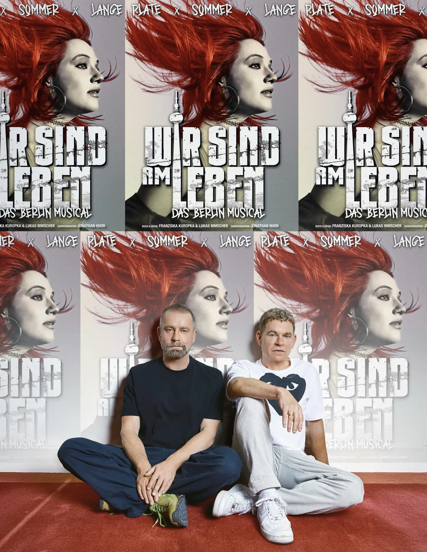 Peter Plate & Ulf Leo Sommer sitzen vor einer Wand mit Postern des Musicals 'Wir sind am Leben' mit einer Frau mit rotem Haar und dem Berliner Fernsehturm im Hintergrund.