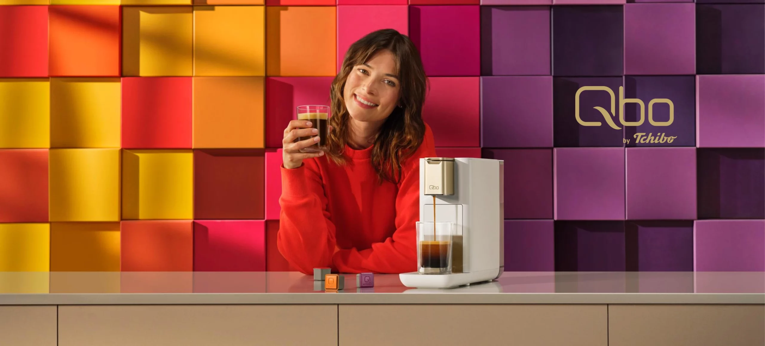 Frau in rotem Pullover sitzt vor einer farbigen Wand aus quadratischen Paneelen und hält ein Glas, daneben steht eine weiße Kaffeemaschine mit Kaffeetasse und Kapseln auf der Theke, links oben das Logo 'Qbo by Tchibo'.