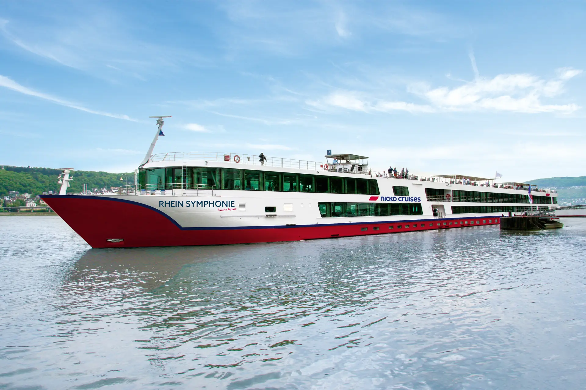 Die RHEIN SYMPHONIE liegt am Anleger. Kiel und Bug sind rot gestrichen. Dicht über der Wasseroberfläche wechselt die Farbe zu Weiß. Große Glasfenster um das Schiff herum lassen das Innere erahnen, ohne das Details erkennbar sind. Menschen stehen an Deck. An der Schiffsseite steht in blauen Buchstaben der Name des Schiffes und der der Reederei nicko cruises.
