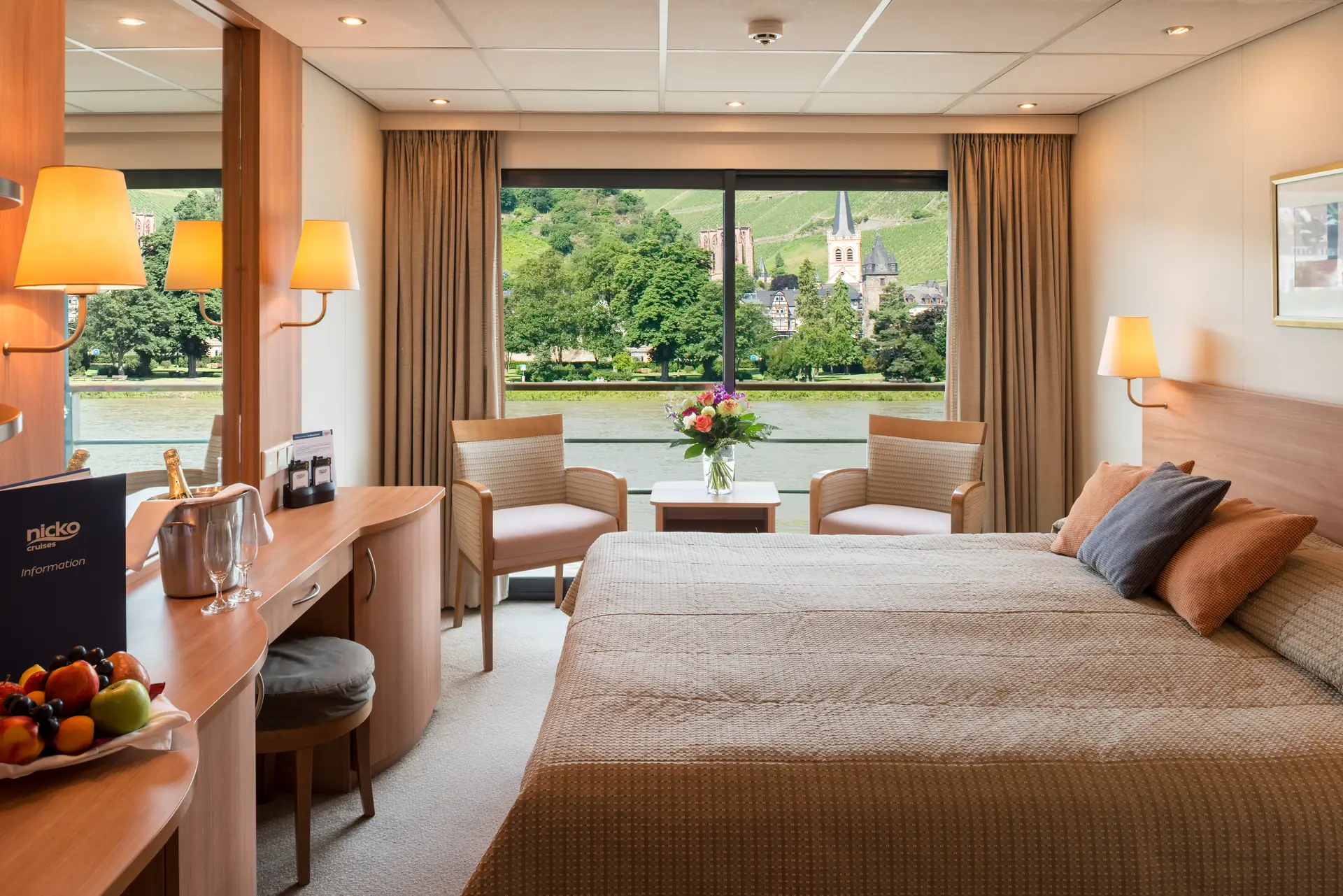 2-Bett-Kabine Oberdeck auf der RHEIN SYMPHONIE. Beige Stoffe und Teppich, zusammen mit Holzelementen an allen Möbeln erzeugen mit dem warmen Licht der Wandlampen eine gemütliche Atmosphäre. Draußen vor dem Fenster zieht eine Stadt mit Kirchturm vorbei.