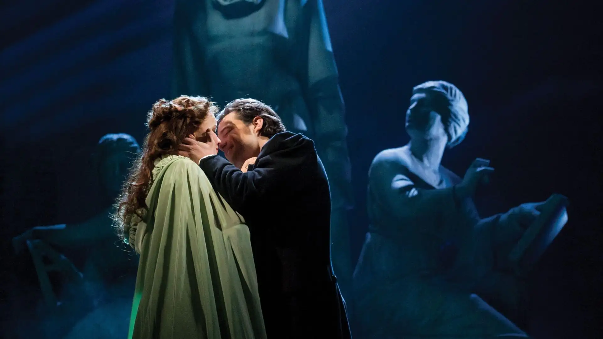 Zwei Personen in einer Bühnenaufnahme des Musicals Phantom der Oper, eine Person hält die andere zärtlich am Gesicht, im Hintergrund sind verschwommene Statuen oder Figuren zu sehen.