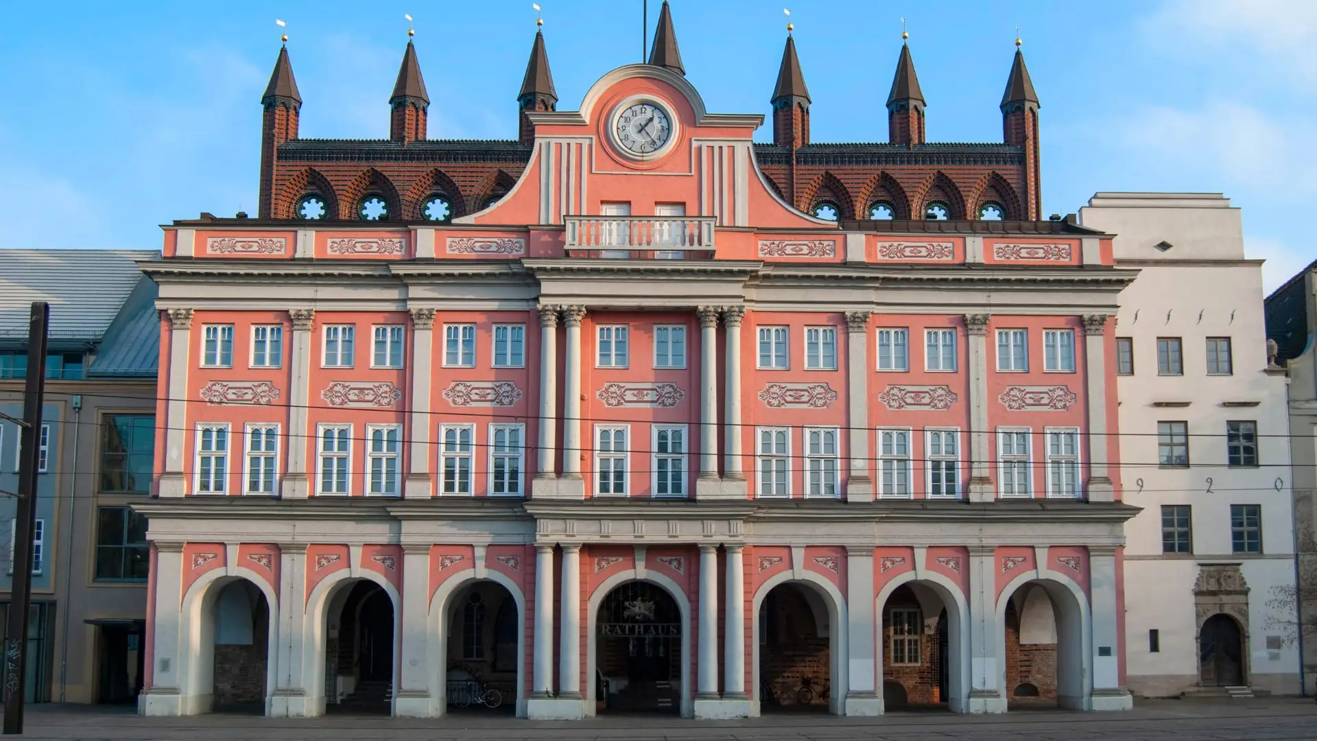 Rathaus Rostock mit rosa Fassade, mehreren Bögen im Erdgeschoss, Uhr und spitzen Türmchen auf dem Dach.