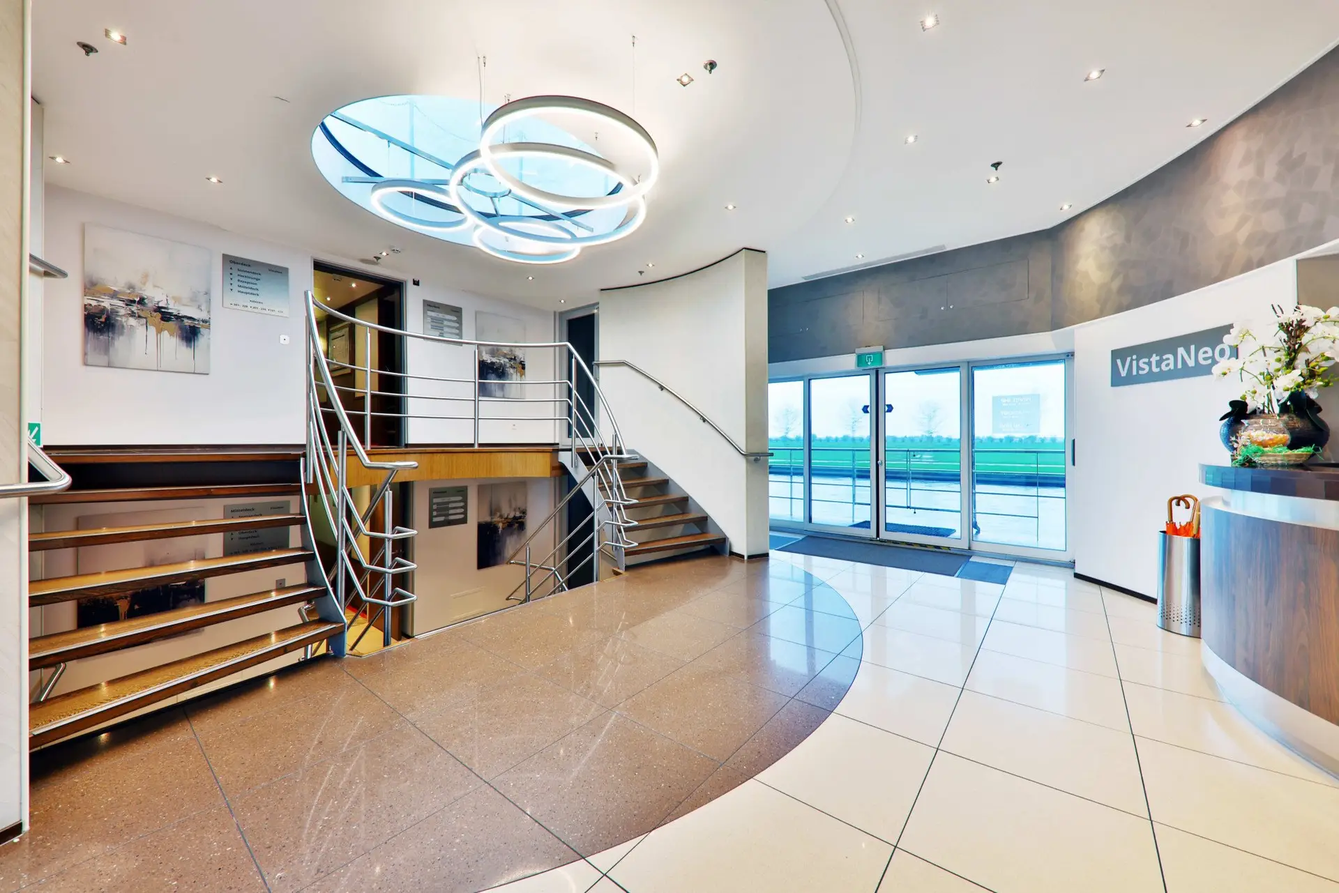 Heller Empfangsbereich mit geschwungener Treppe, runden Deckenleuchten und Glas-Eingangstür mit 'VistaNeon'-Schild