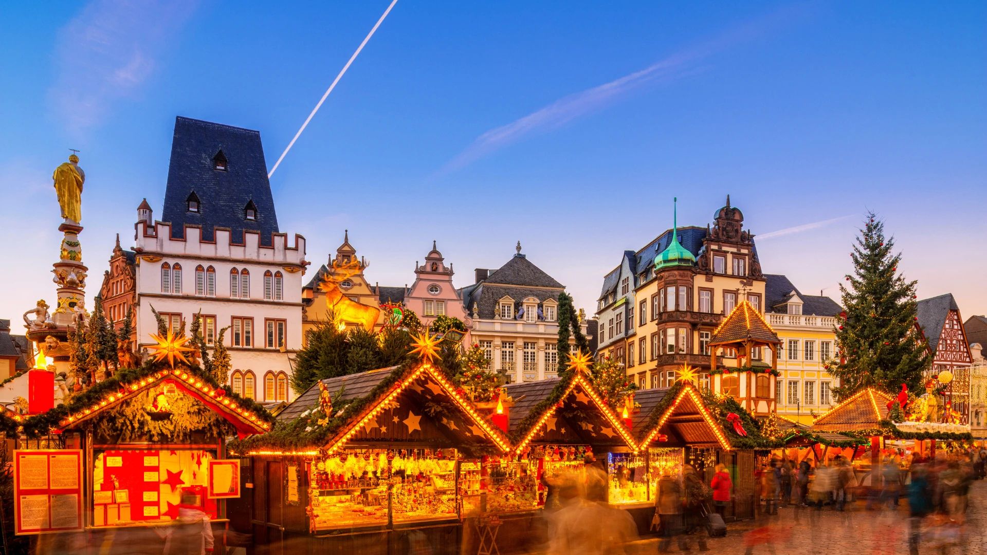 Weihnachtsmarkt mit beleuchteten Holzhütten vor historischen Gebäuden und einem großen Weihnachtsbaum in Trier bei Abendlicht