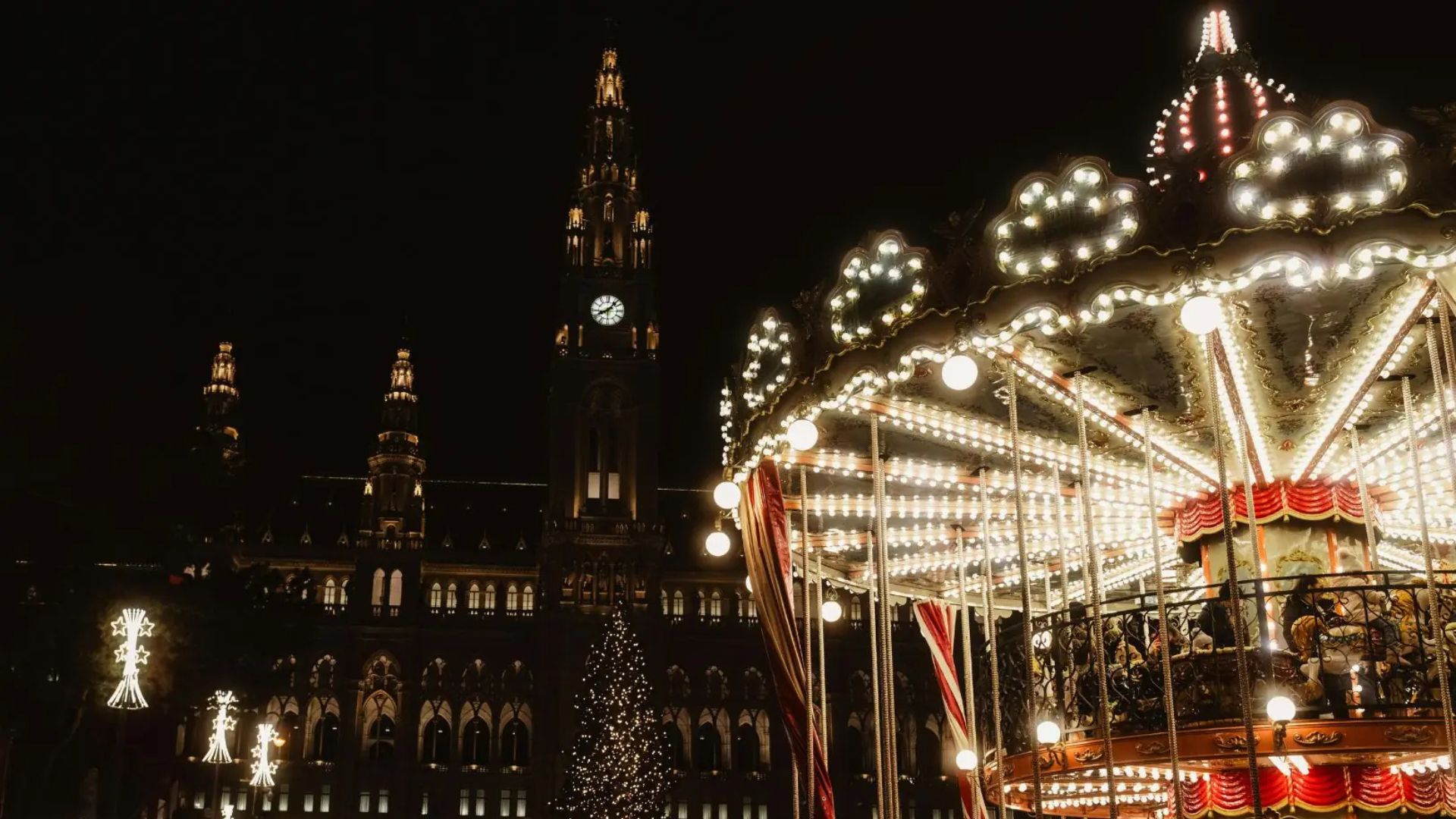 Beleuchtetes Karussell im Vordergrund, im Hintergrund das Rathaus von Wien mit Uhr und Weihnachtsbaum. Nachtaufnahme.