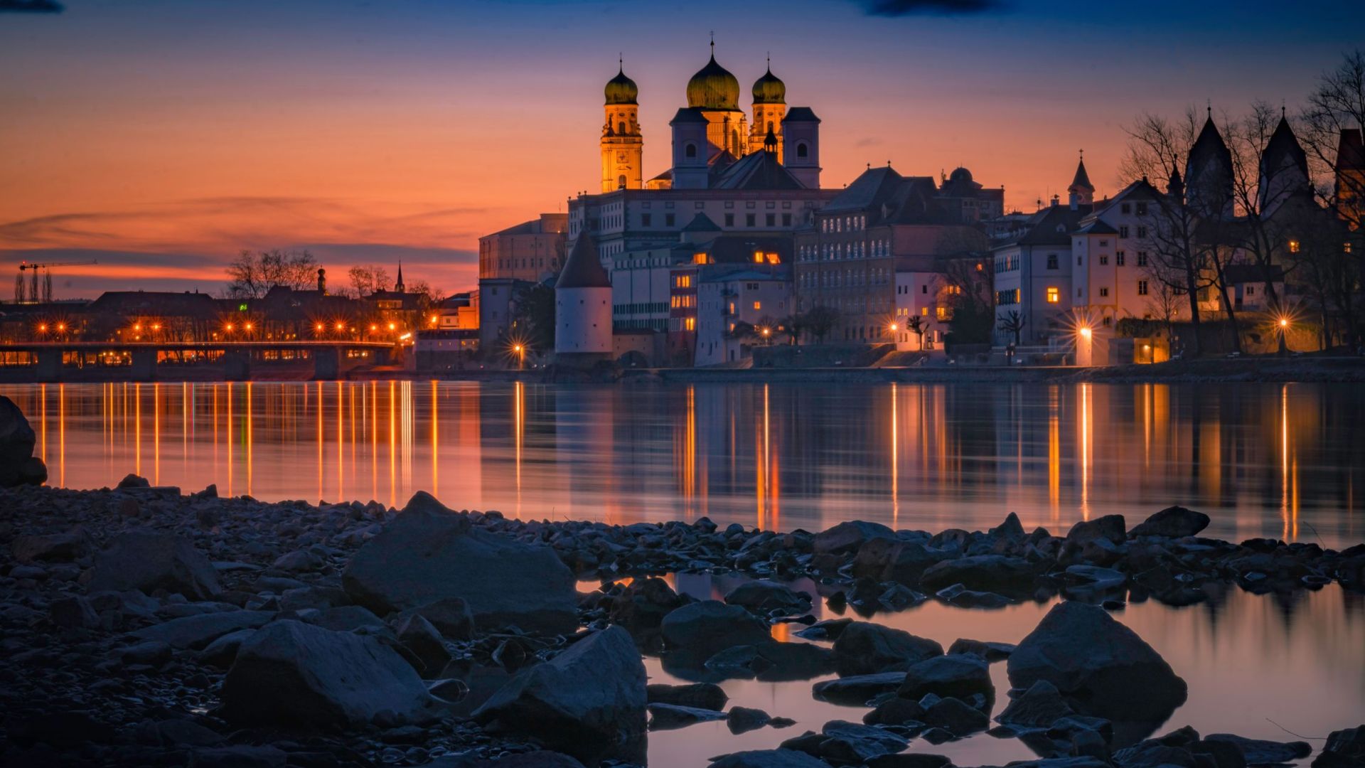 Stadtansicht bei Sonnenuntergang mit reflektierenden Lichtern im Wasser, Felsen im Vordergrund, historische Gebäude mit Kuppeln im Hintergrund.