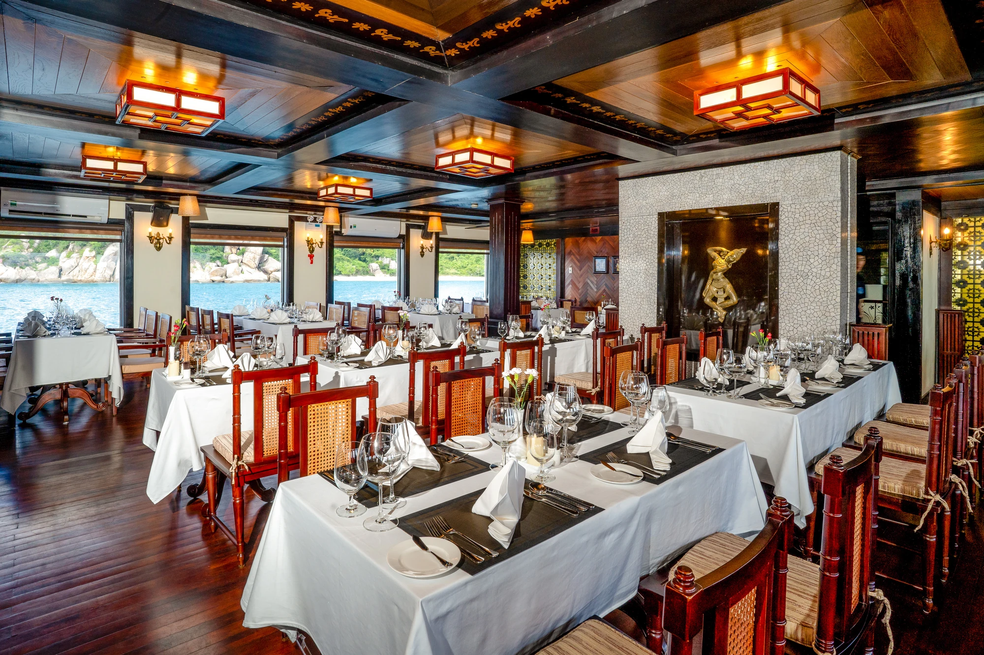 Elegantes Restaurant mit Holztischen und weißen Tischdecken. Stühle aus Holz mit Rattan. Gläser und Besteck auf jedem Tisch. Beleuchtung durch rote Lampen.