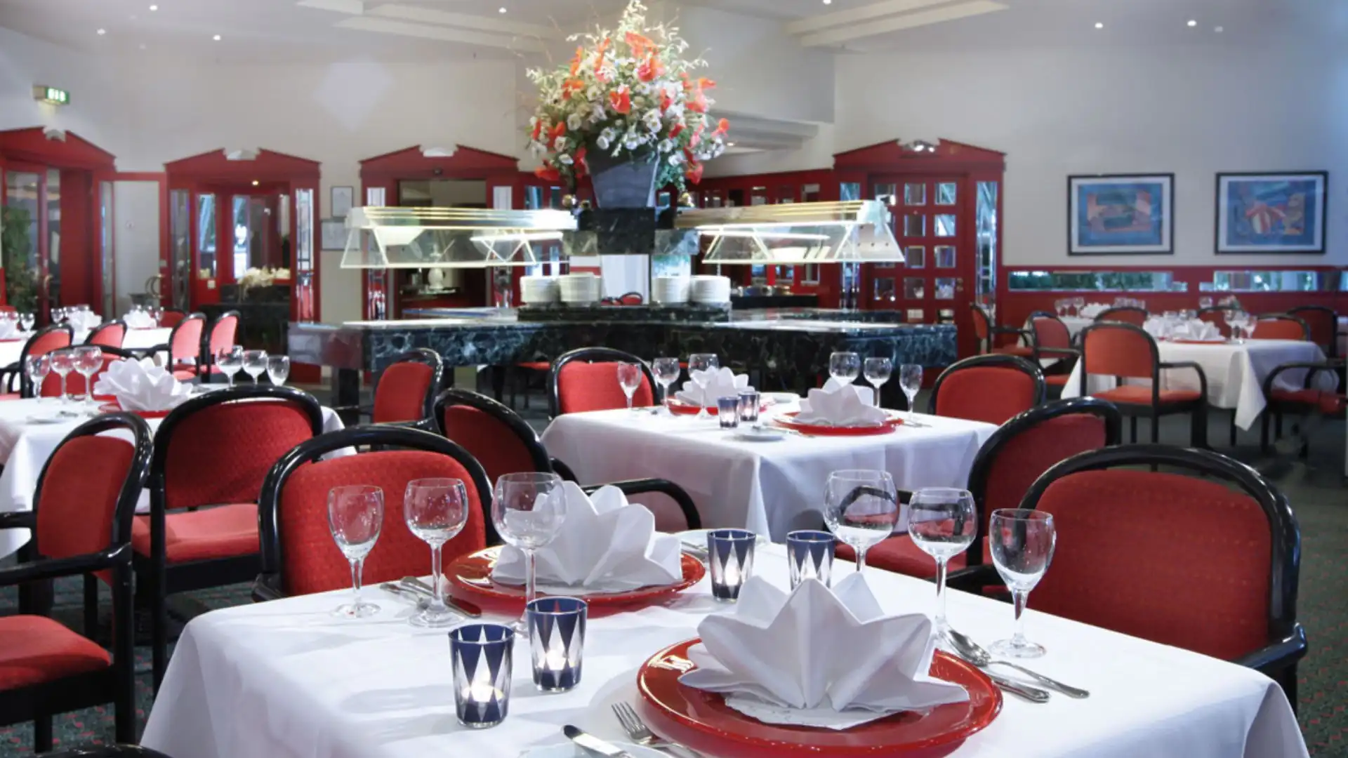 Elegantes Restaurant mit rot gepolsterten Stühlen, weiß gedeckten Tischen und einem großen Blumenarrangement auf einem zentralen Buffet im Hotel Kaiserhof Landshut