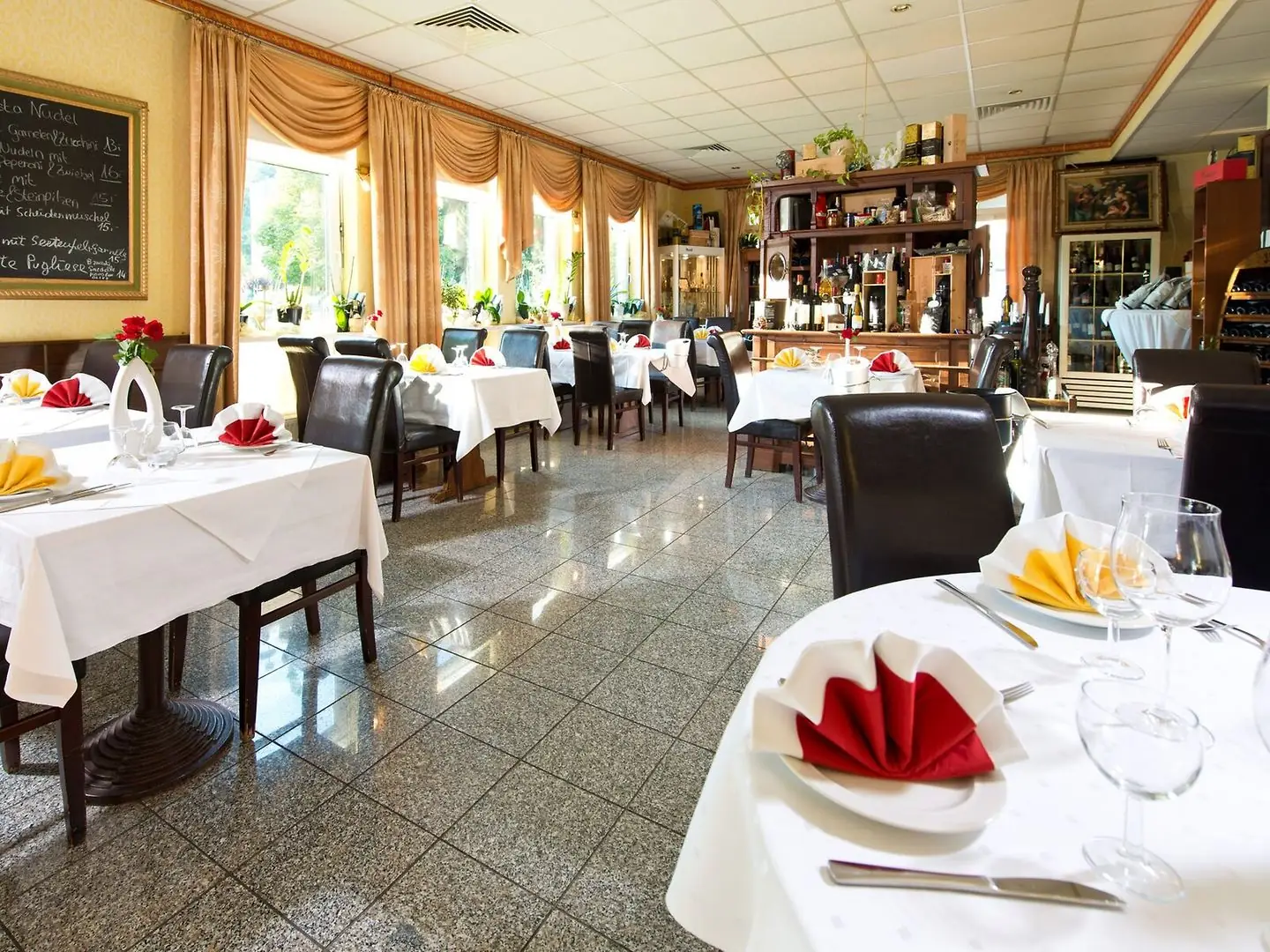 Helles Restaurant mit weißen Tischdecken, gefalteten Servietten in Rot und Gelb, dunklen Stühlen und großen Fenstern mit Vorhängen im ACHAT Hotel Monheim am Rhein