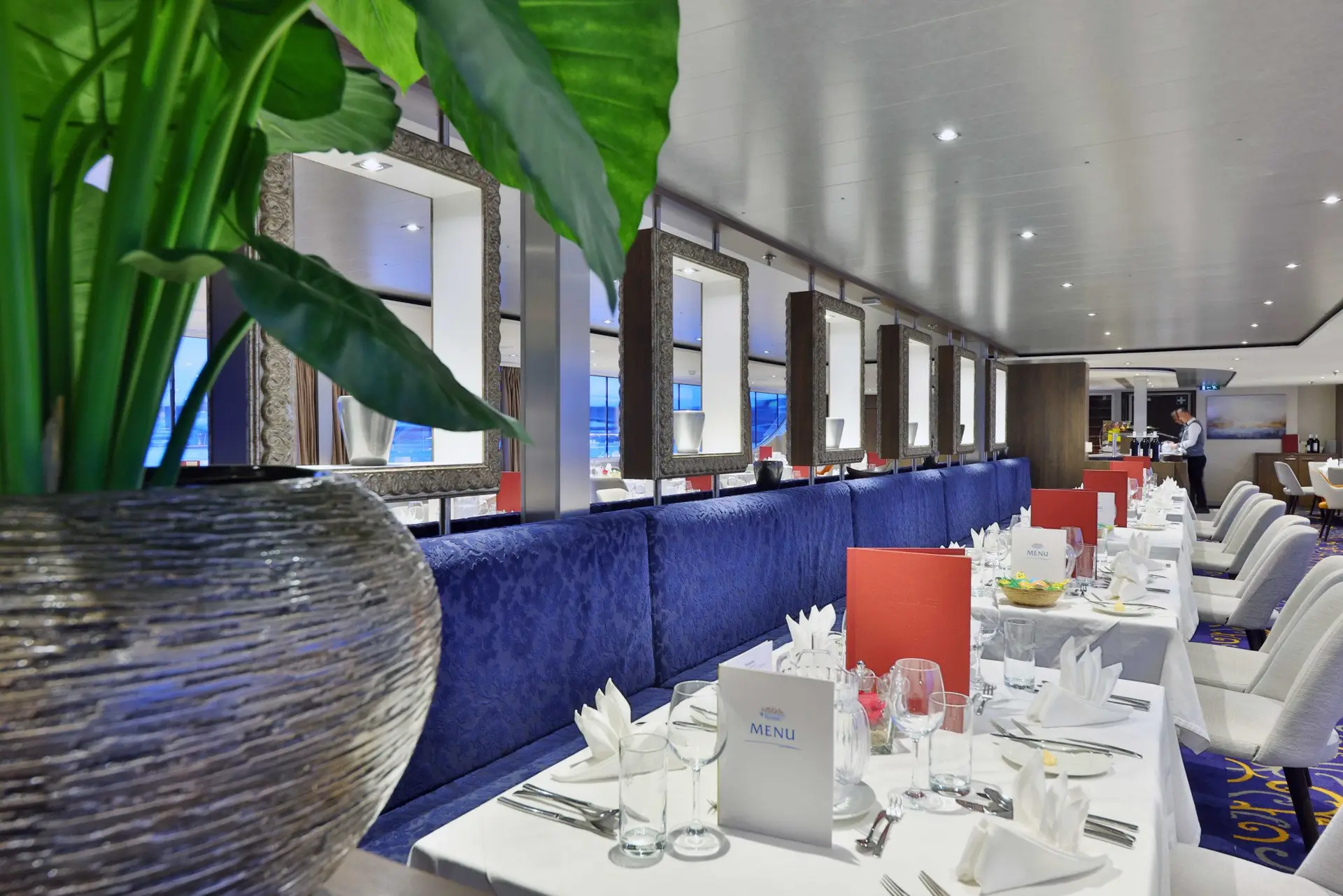 Restaurant mit gedeckten Tischen, blauer Polsterbank, weißen Stühlen und großen Spiegeln an der Wand