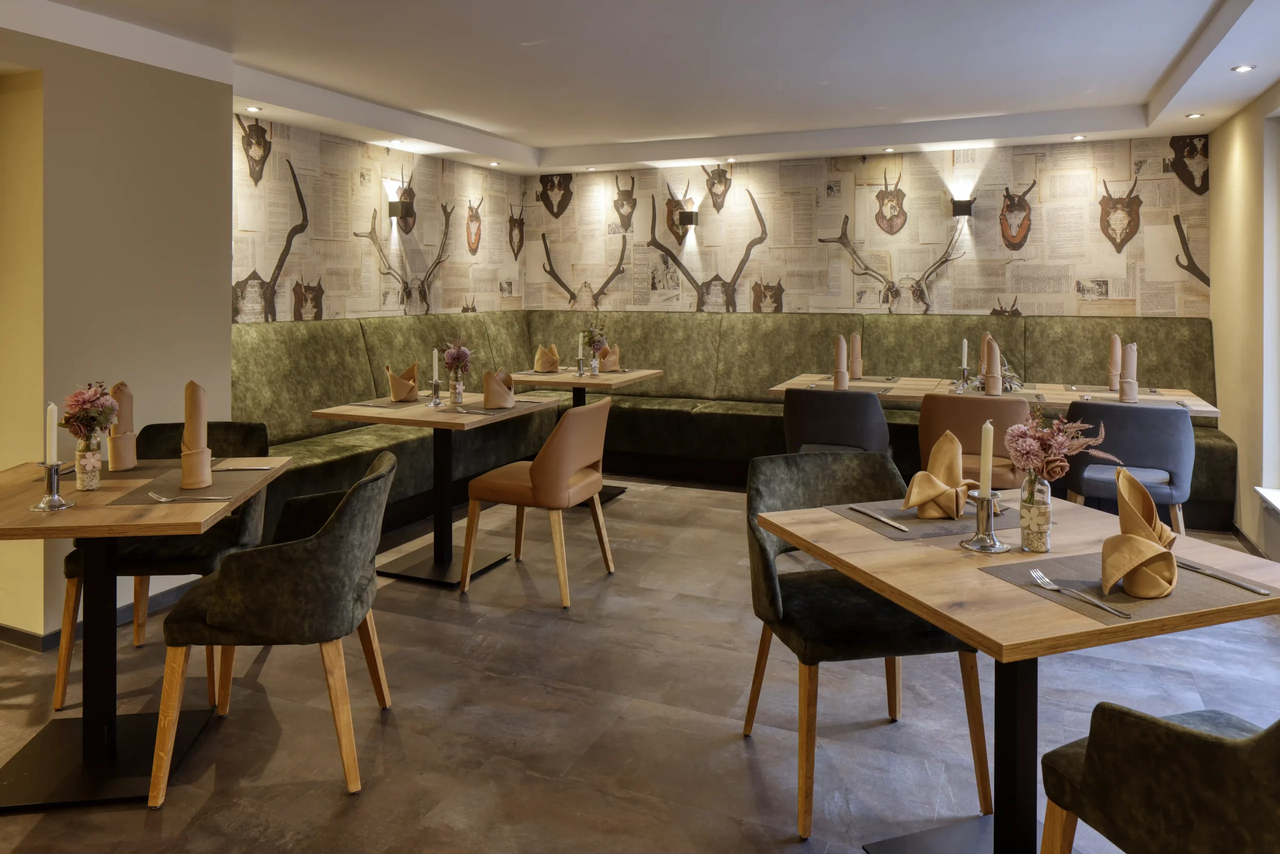 Gemütlicher Gastraum im Restaurant Waldzimmer mit grüner Polsterbank, Holztischen, verschiedenen Stühlen und einer Wand mit Hirschgeweihen als Dekoration.