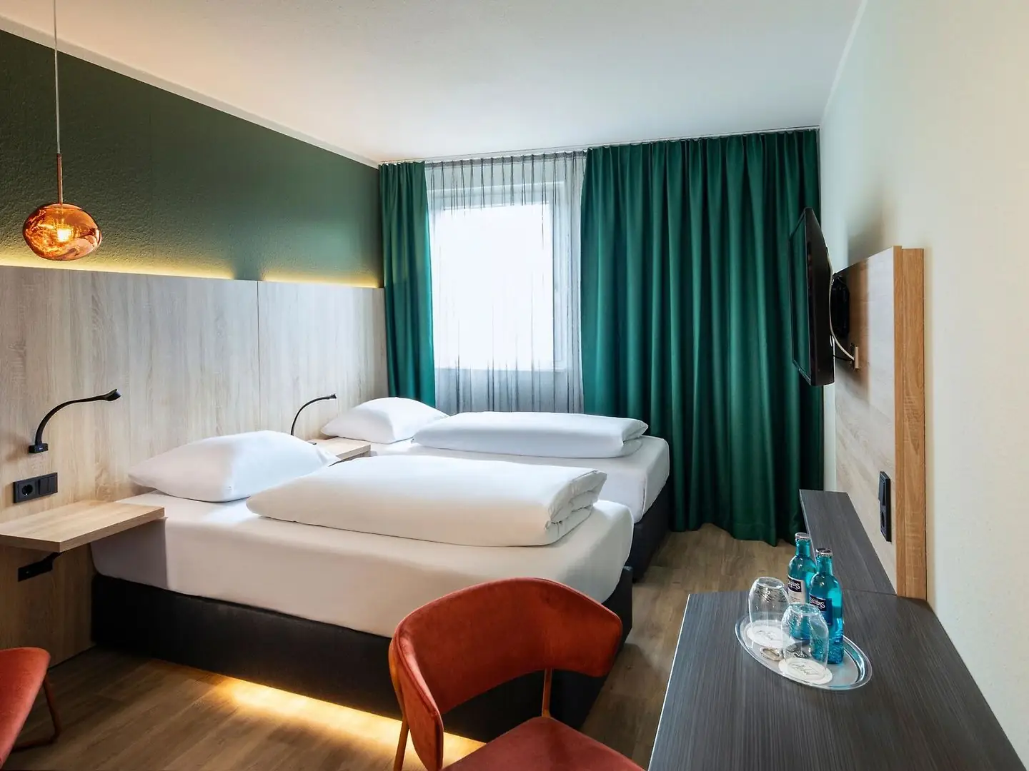 Hotelzimmer mit zwei Einzelbetten, Nachttischlampen, grünem Vorhang, Wandfernseher und zwei roten Stühlen