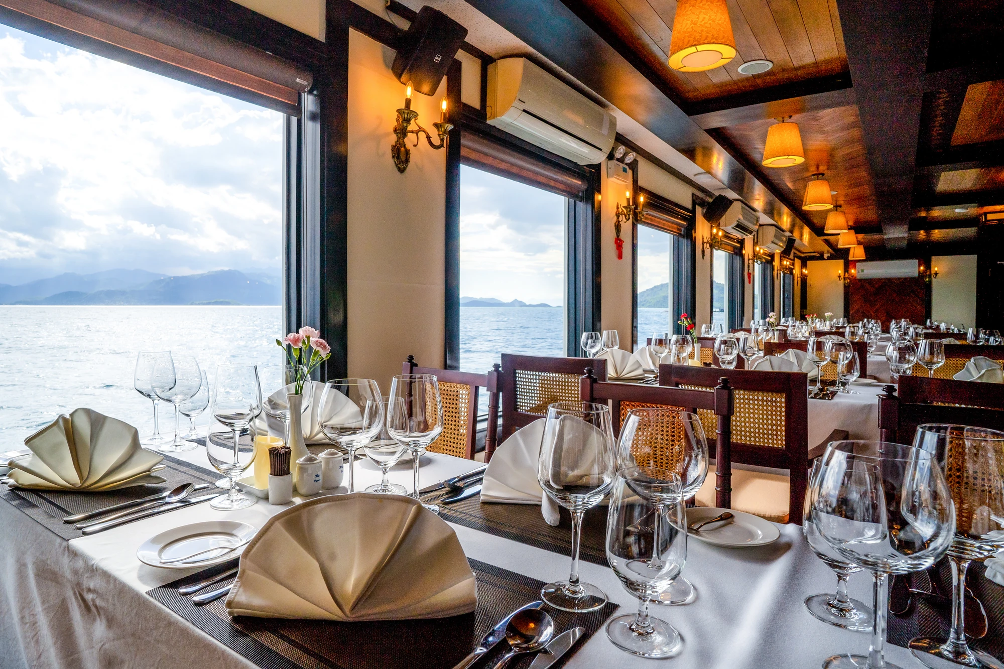 Elegante Restaurantansicht mit gedeckten Tischen, Gläsern und Servietten. Große Fenster mit Blick auf das Wasser und die Landschaft im Hintergrund.