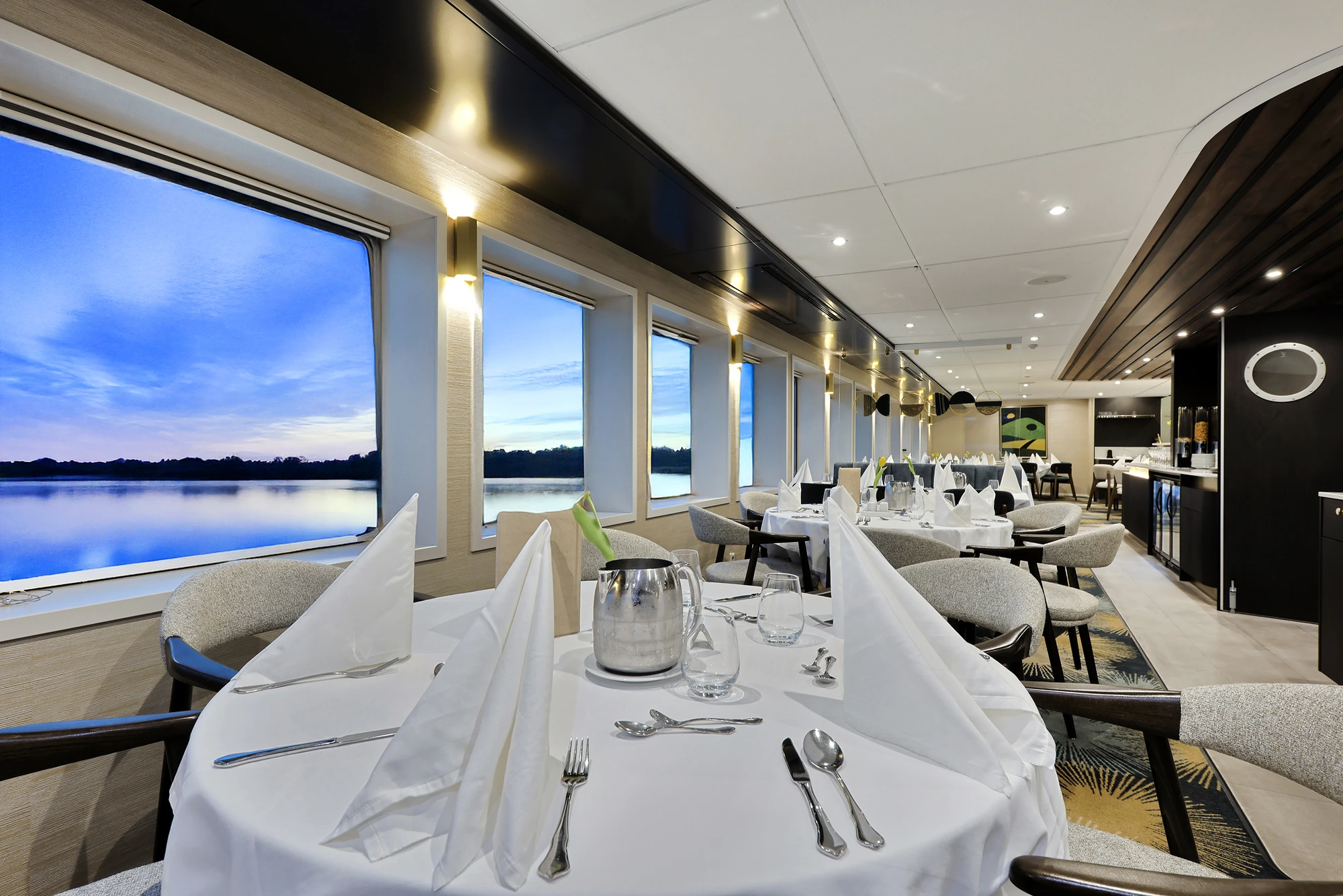 Elegantes Restaurant mit gedeckten Tischen und großen Fenstern mit Blick auf einen See bei Abenddämmerung