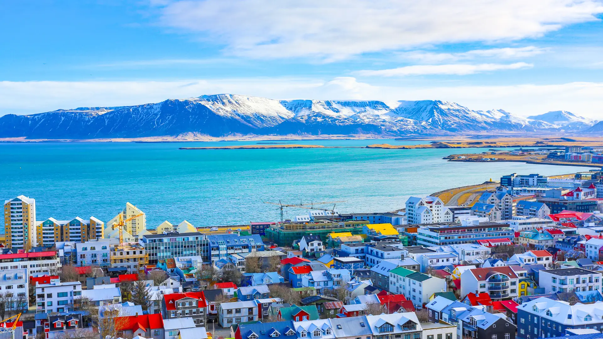Blick auf Reykjavik mit bunten Häusern am Wasser und schneebedeckten Bergen im Hintergrund unter blauem Himmel.