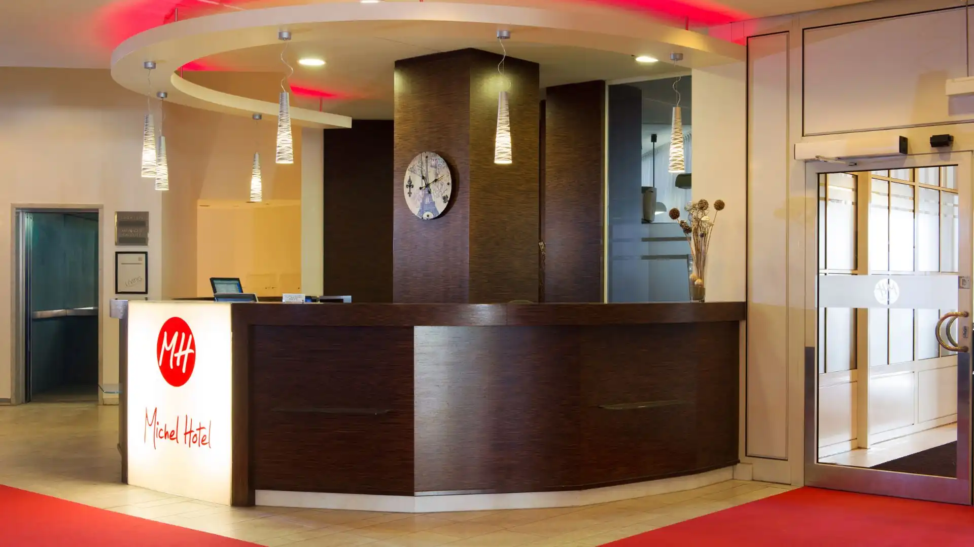 Rezeption des Hotel Kaiserhof Landshut mit dunklem Empfangstresen, beleuchtetem Logo 'MH Michel Hotel', rundem Deckenlicht und Glastür zum Eingangsbereich.