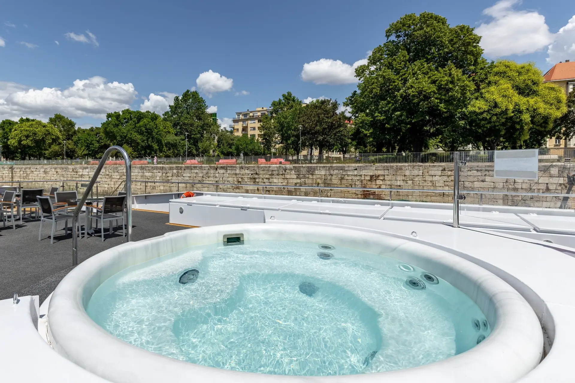 Runder Whirlpool mit sprudelndem Wasser auf einer Terrasse, im Hintergrund Bäume und Gebäude unter blauem Himmel mit Wolken.