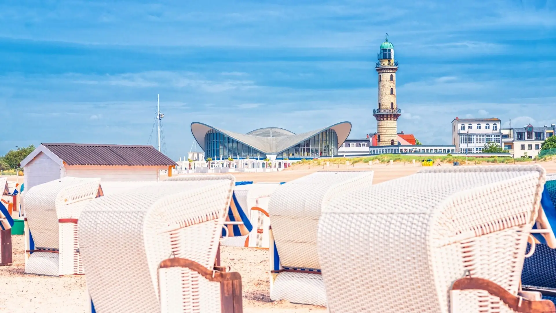 Strandkörbe am Sandstrand in Rostock mit Leuchtturm und moderner Architektur im Hintergrund.