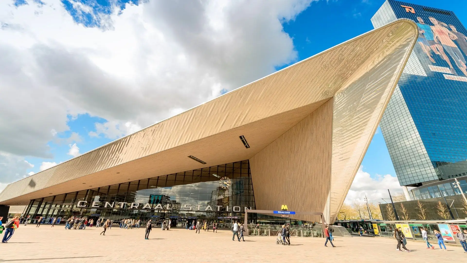 Außenansicht des modernen Gebäudes Rotterdam Centraal mit markantem, spitz zulaufendem Dach und Glasfassade, davor Menschen auf dem Platz.