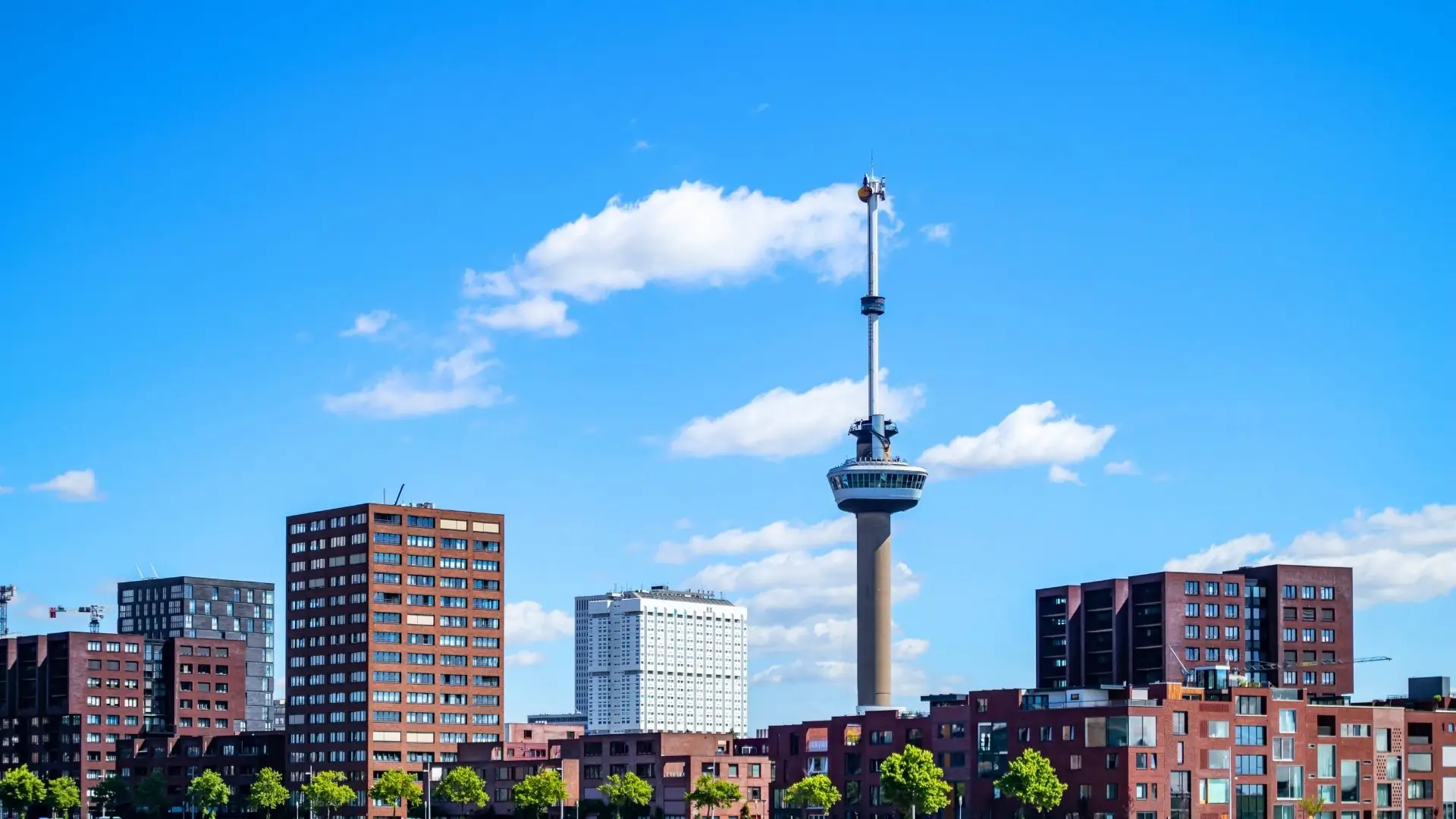 Euromast-Turm in Rotterdam vor blauem Himmel mit einzelnen Wolken, umgeben von mehrstöckigen Wohn- und Bürogebäuden.
