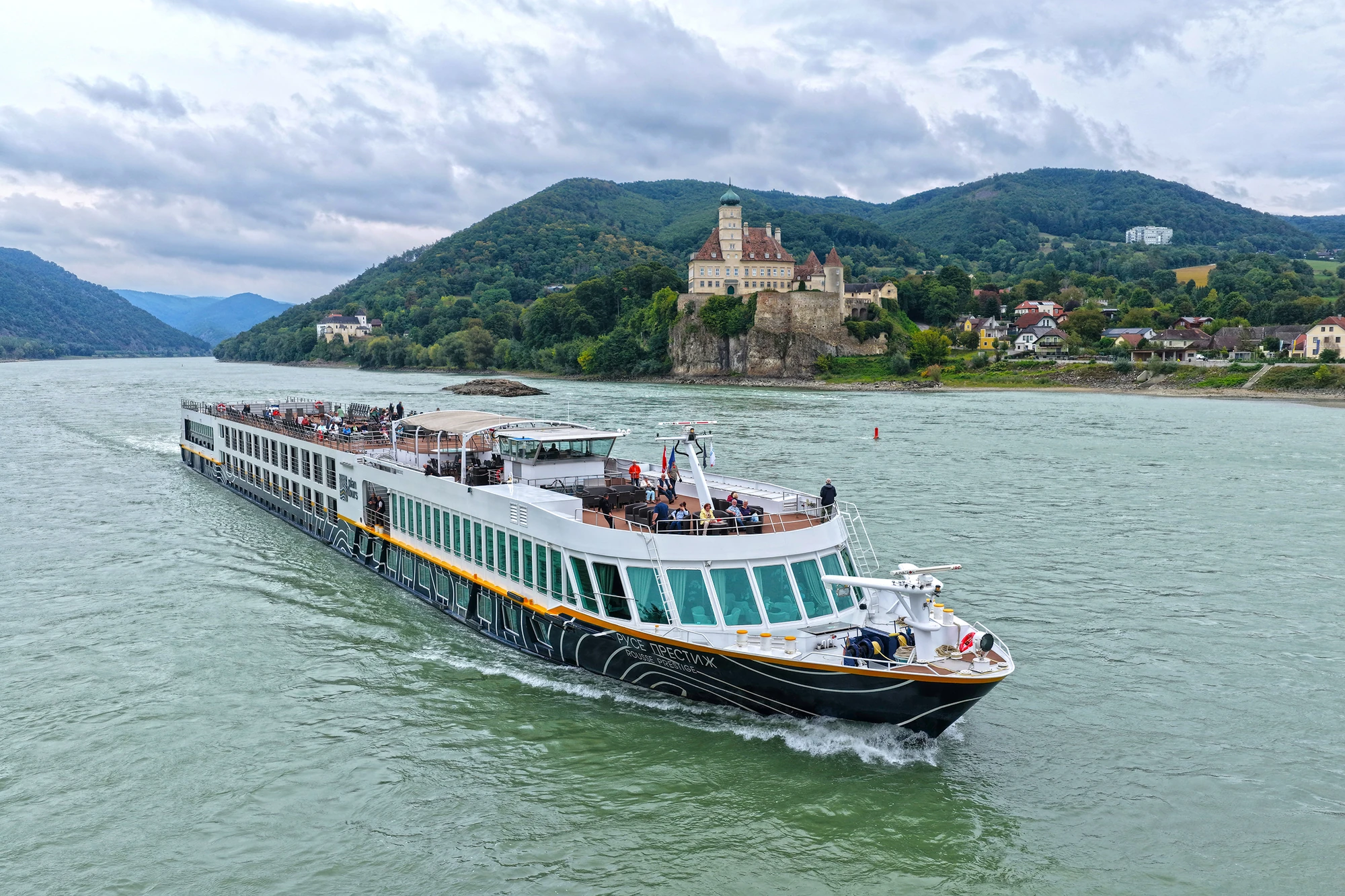 Flusskreuzfahrtschiff fährt auf einem breiten Fluss mit bewaldeten Hügeln und einem Schloss im Hintergrund