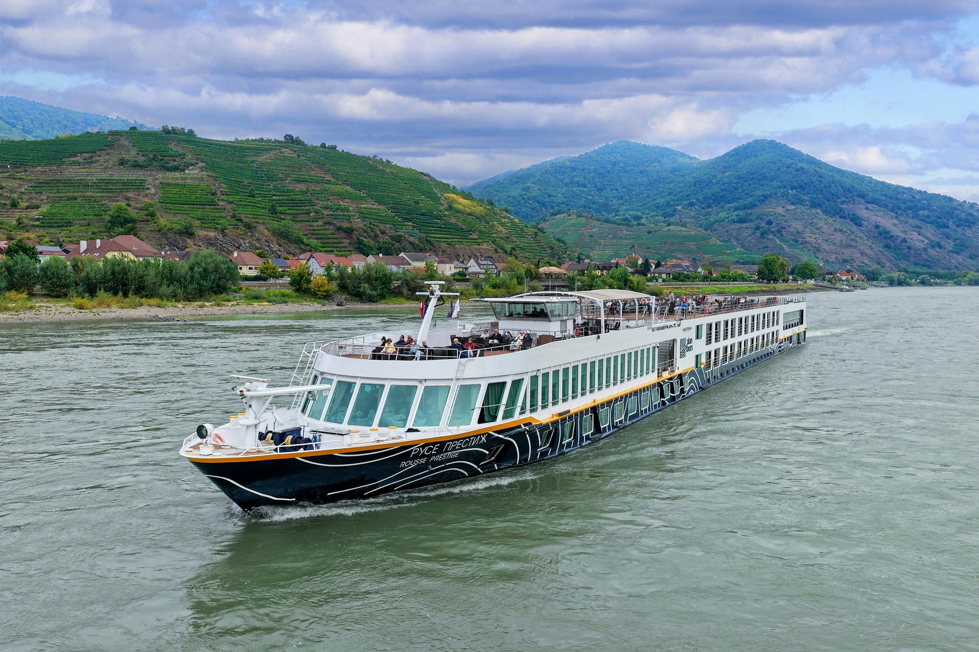 Flusskreuzfahrtschiff mit Passagieren auf dem Oberdeck fährt auf einem Fluss vor bewaldeten und terrassierten Hügeln