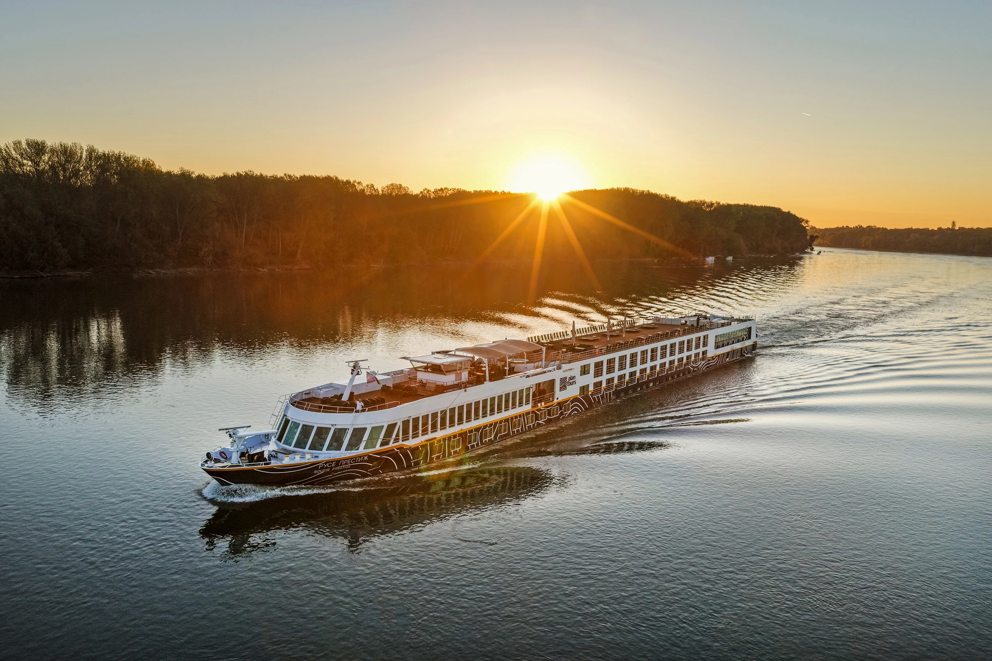 Flusskreuzfahrtschiff fährt bei Sonnenaufgang auf einem breiten Fluss mit bewaldeten Ufern