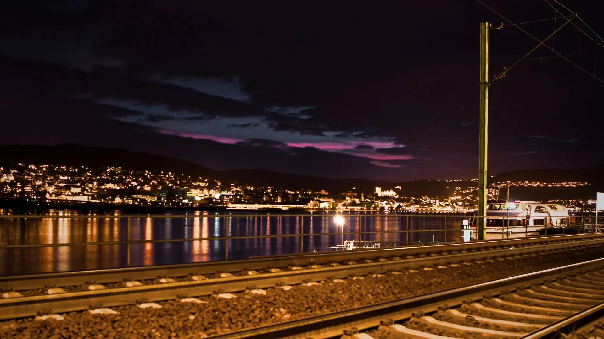 Nachtansicht von Rüdesheim am Rhein mit beleuchteten Gebäuden am Ufer und reflektierenden Lichtern im Wasser. Gleise im Vordergrund.