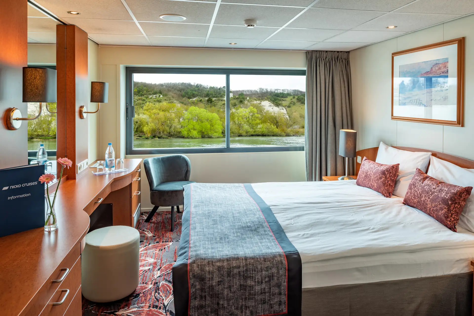 Seine Comtesse, 2-Bett Kabine Deluxe Mitteldeck mit Doppelbett, Schreibtisch, Stuhl und großem Fenster mit Blick auf grüne Landschaft und Wasser.