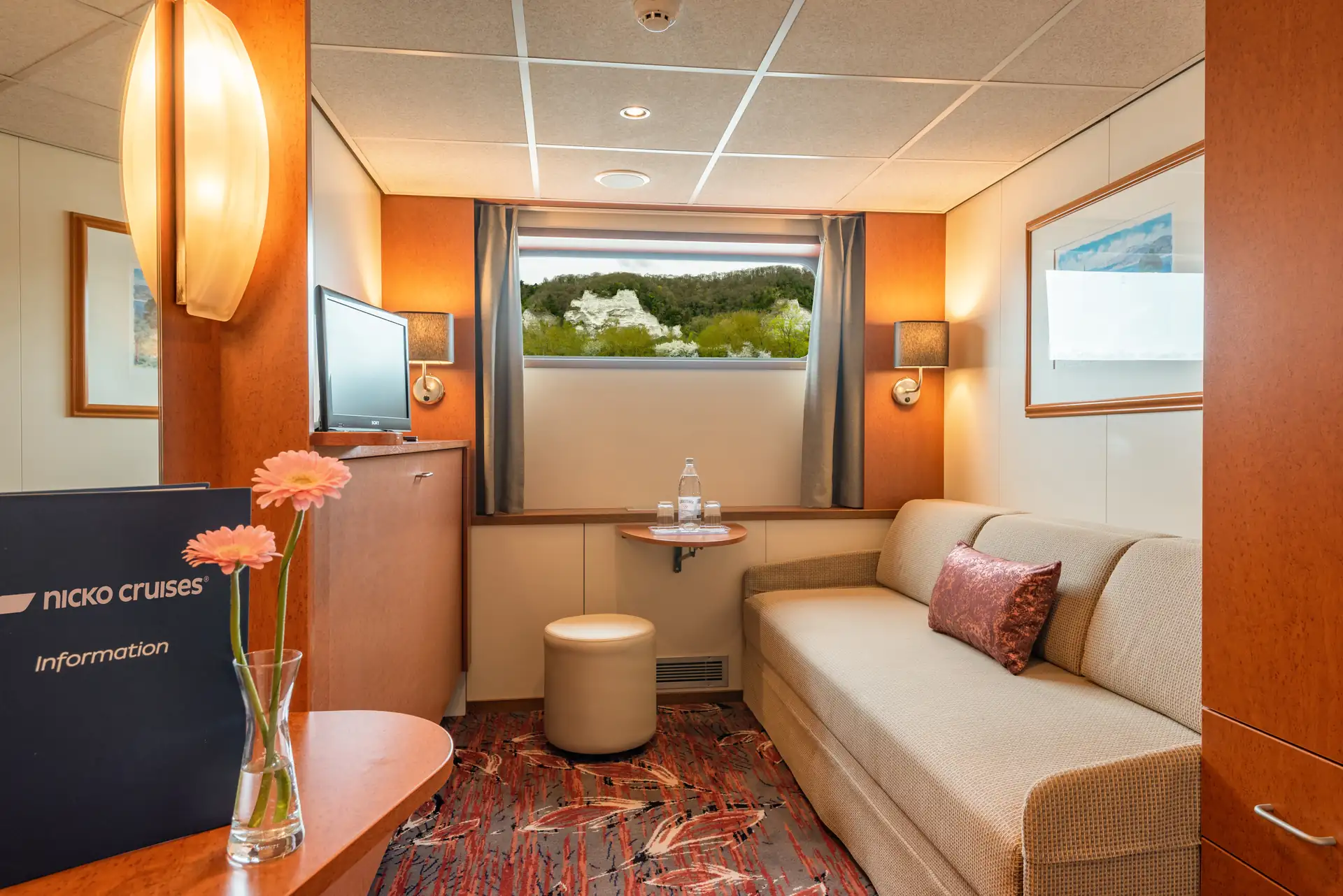 Seine Comtesse, 2-Bett Kabine Hauptdeck mit Sofa, rundem Tisch, Hocker, Fernseher, Fenster mit Blick auf Felsen und Bäume, Vase mit Blumen, Informationsmappe von nicko cruises.