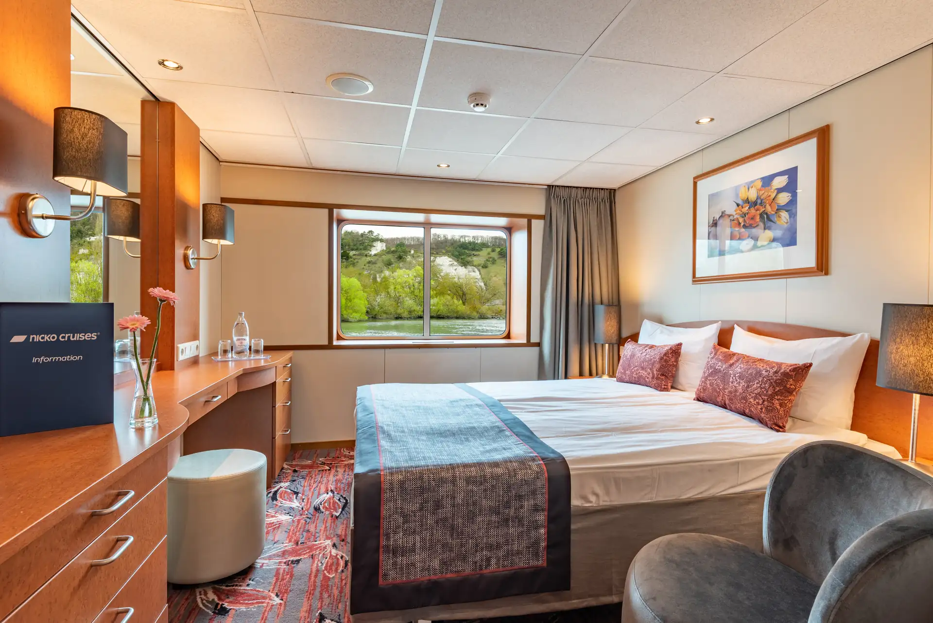 Seine Comtesse, 2-Bett Kabine Mitteldeck mit Doppelbett, zwei Kissen, Schreibtisch mit Stuhl, Fenster mit Blick auf grüne Bäume und Wandbild mit Blumenmotiv