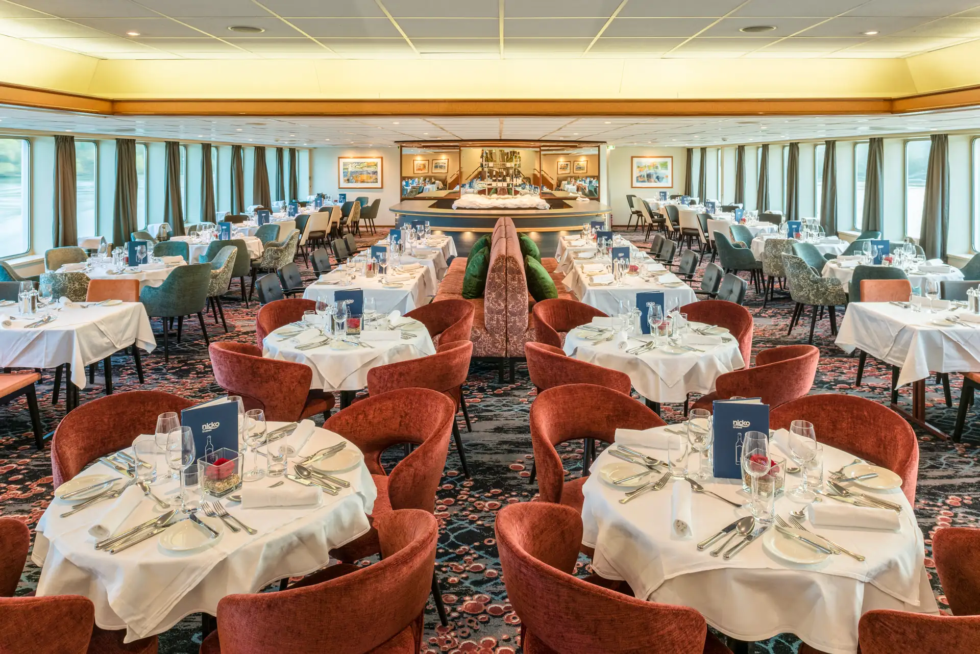 Seine Comtesse Restaurant mit runden Tischen, weiß gedeckt, rot gepolsterte Stühle, Teppichboden und großen Fenstern mit Vorhängen.