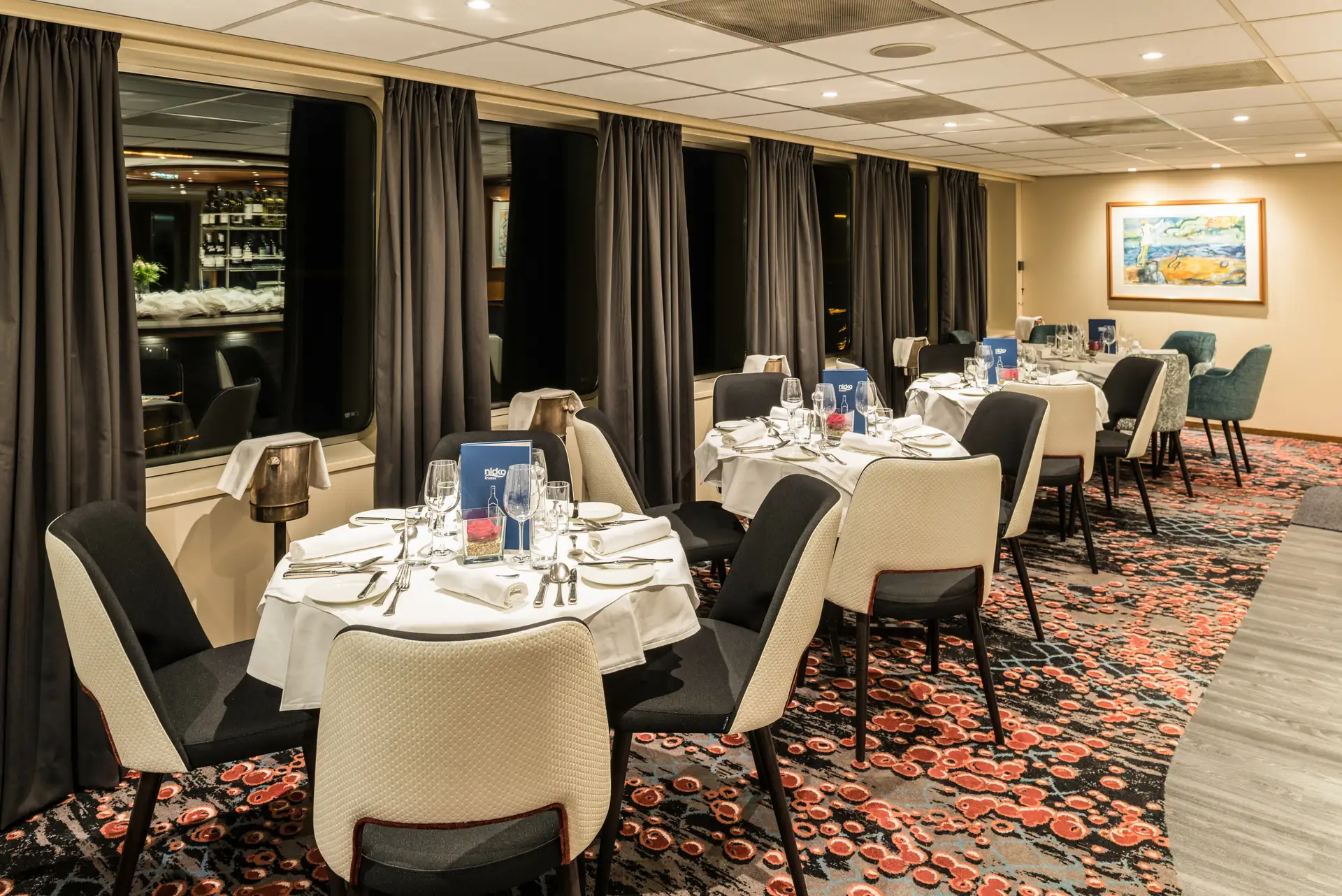 Seine Comtesse Restaurant mit rund gedeckten Tischen, gepolsterten Stühlen, dunklen Vorhängen und Teppich mit rotem Blumenmuster.