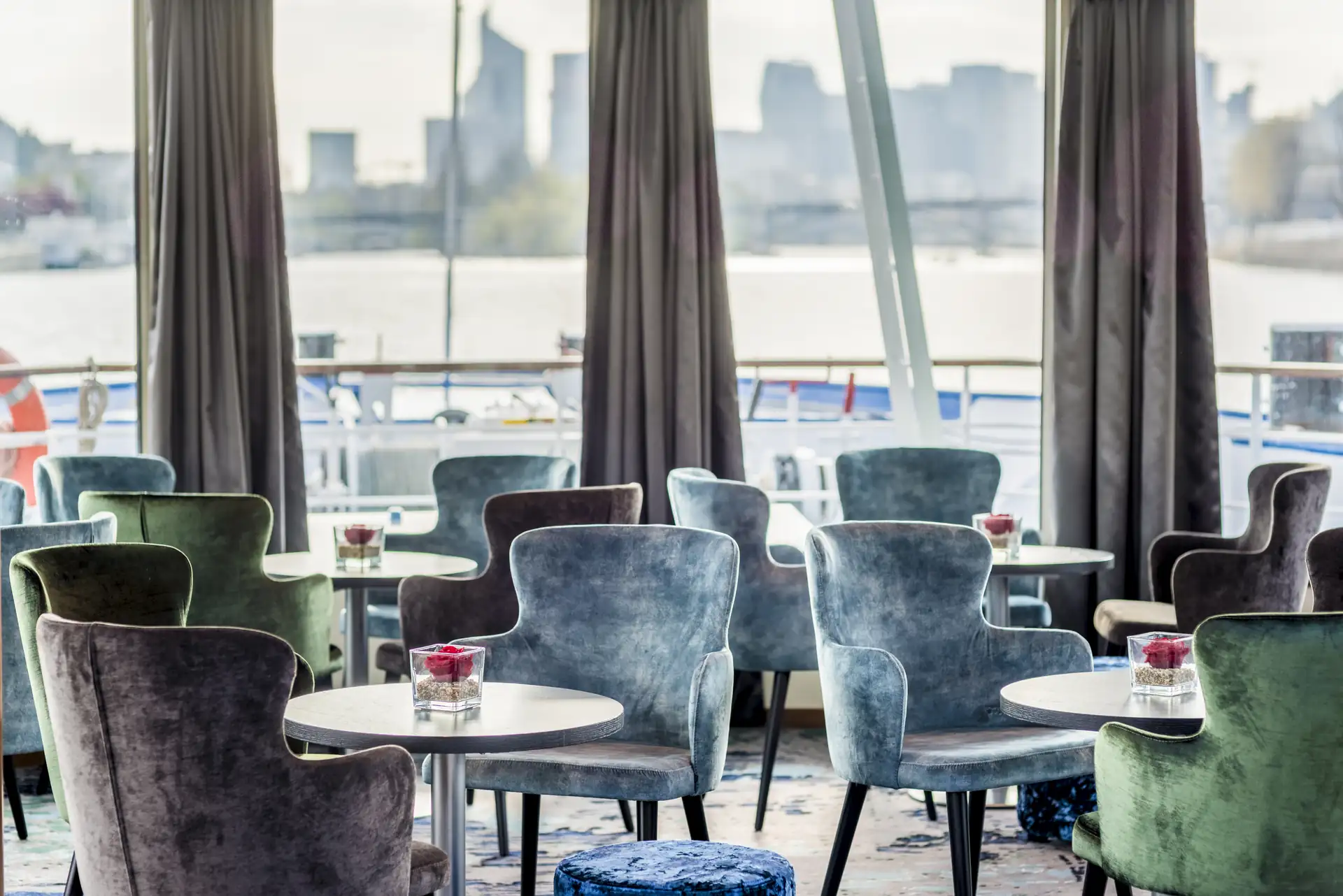 Seine Comtesse Panorama-Salon mit gepolsterten Stühlen in Blau- und Grüntönen und runden Tischen vor großen Fenstern mit Blick auf Wasser und Stadt.