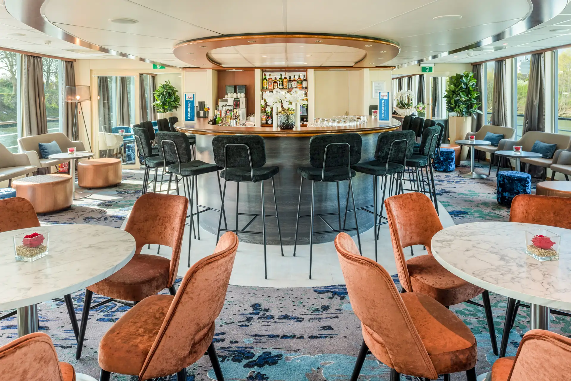 Seine Comtesse Bar mit schwarzer Theke und hohen Barhockern, umgeben von Tischen mit braunen Stühlen und Sofas in einem hellen Raum mit großen Fenstern.