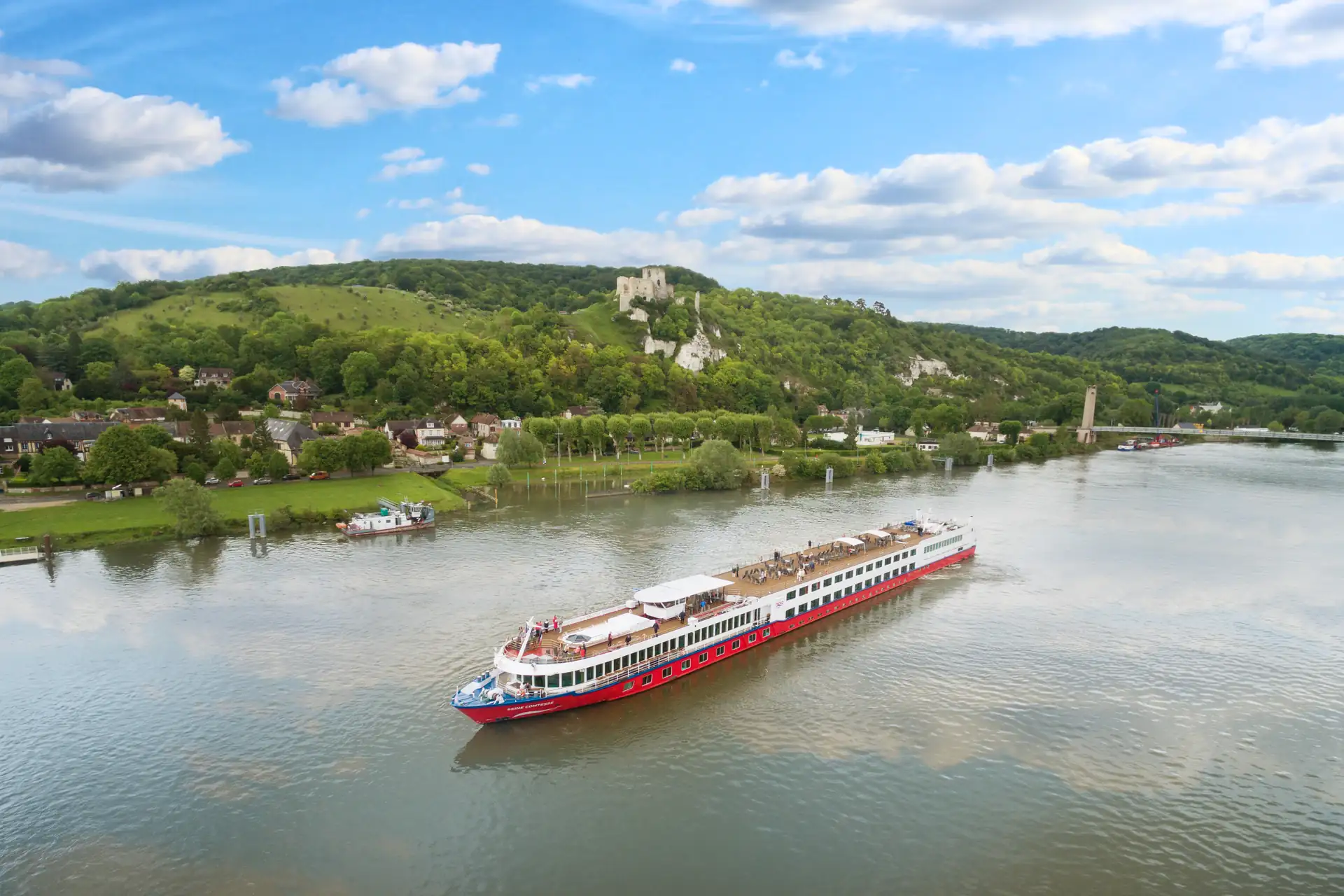 Seine Comtesse mit rotem Rumpf auf ruhigem Fluss vor bewaldetem Hügel mit Burgruine und Dorf unter blauem Himmel mit Wolken.
